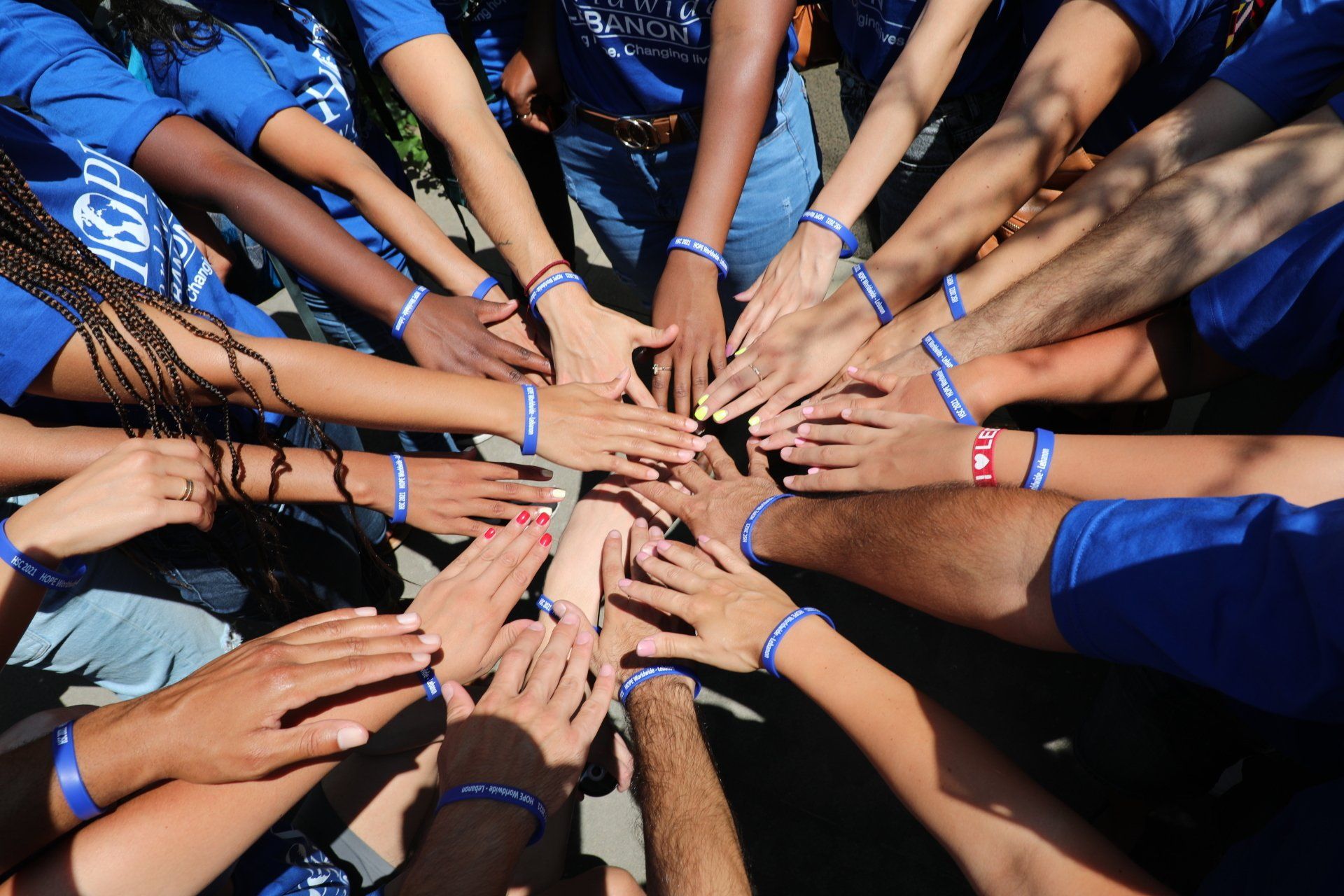 Many hands stacked together outdoors, wearing blue shirts and colorful wristbands