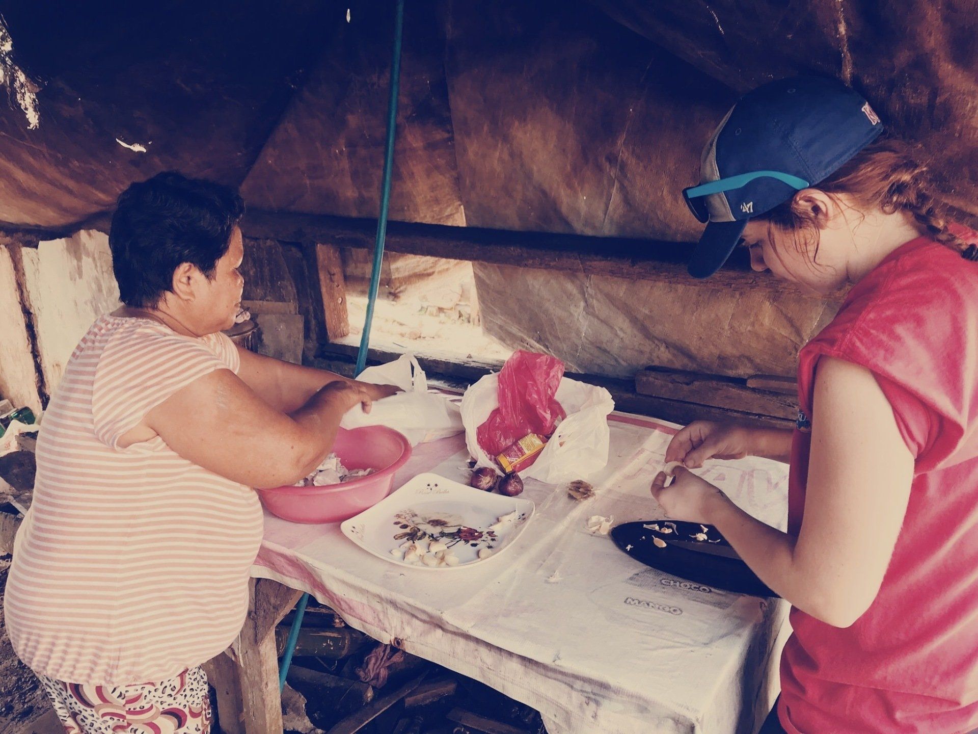 Two people preparing food at a table under a rustic shelter, with bowls, plates, and ingredients spread out.