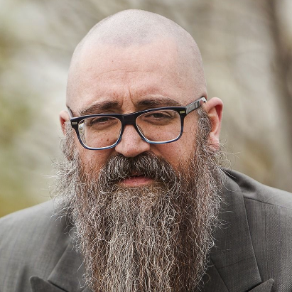 Close-up of a bald man with glasses and a long gray beard wearing a suit outdoors