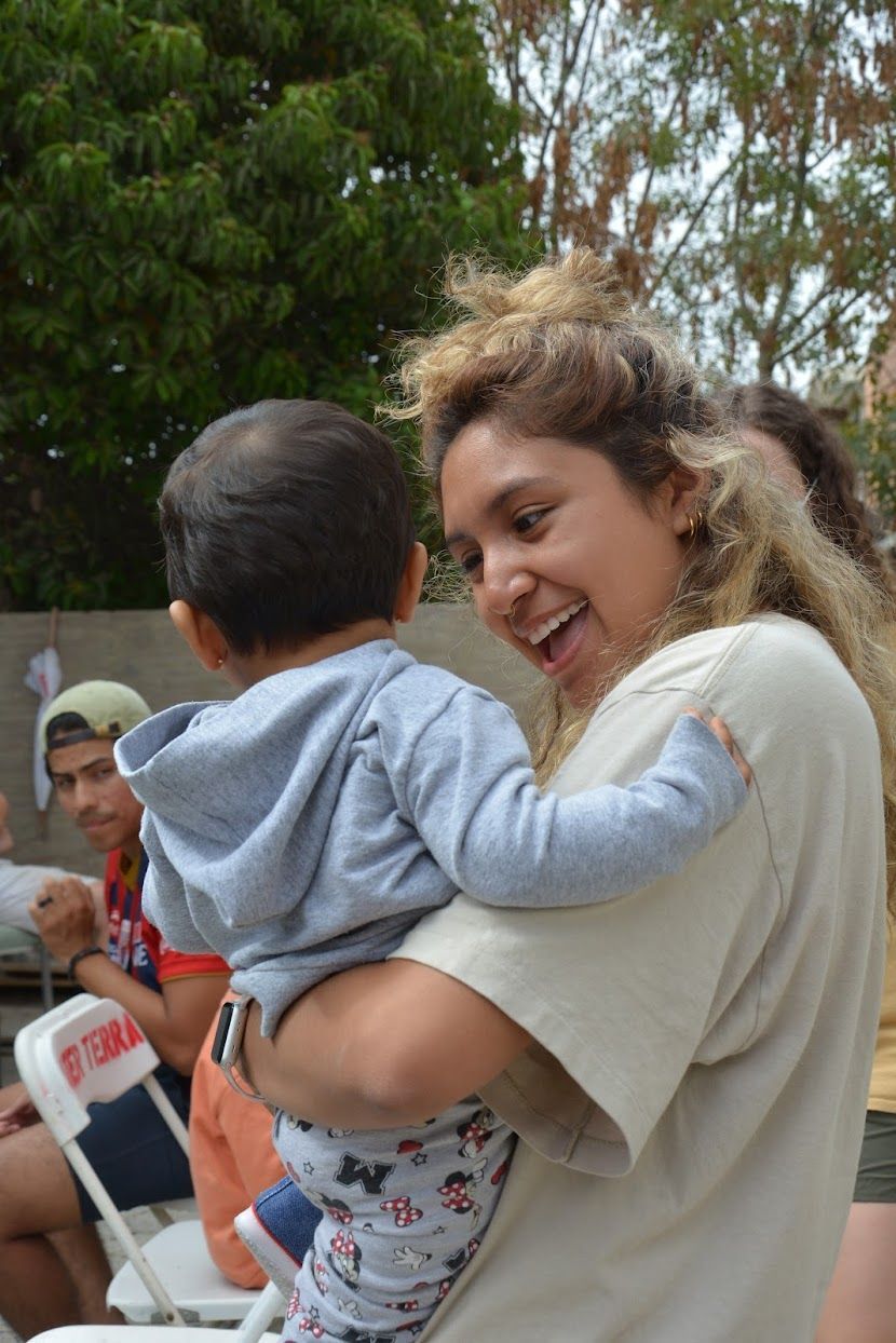 Woman smiling and holding a baby outdoors, with trees in the background