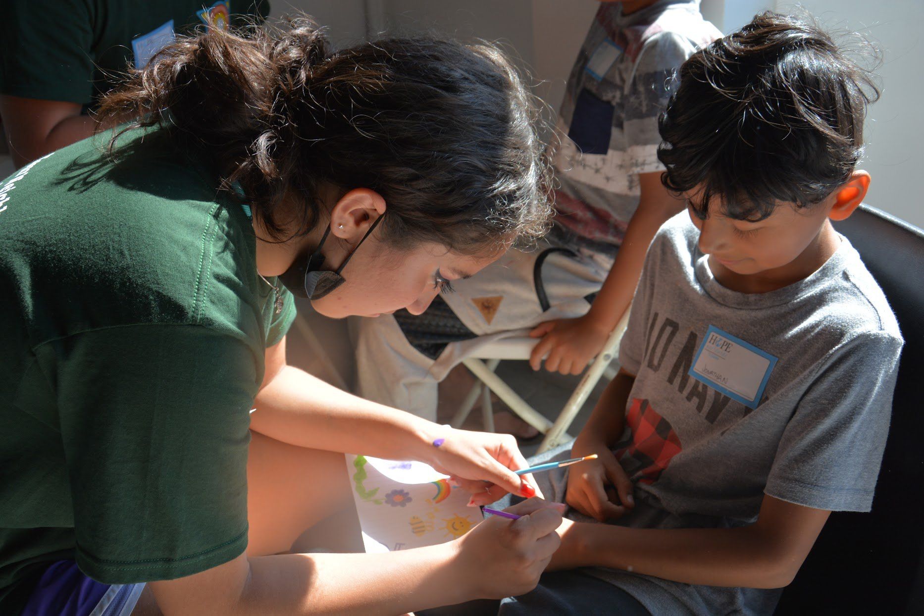 Woman in green shirt helps a child cut paper at a table in a bright room.