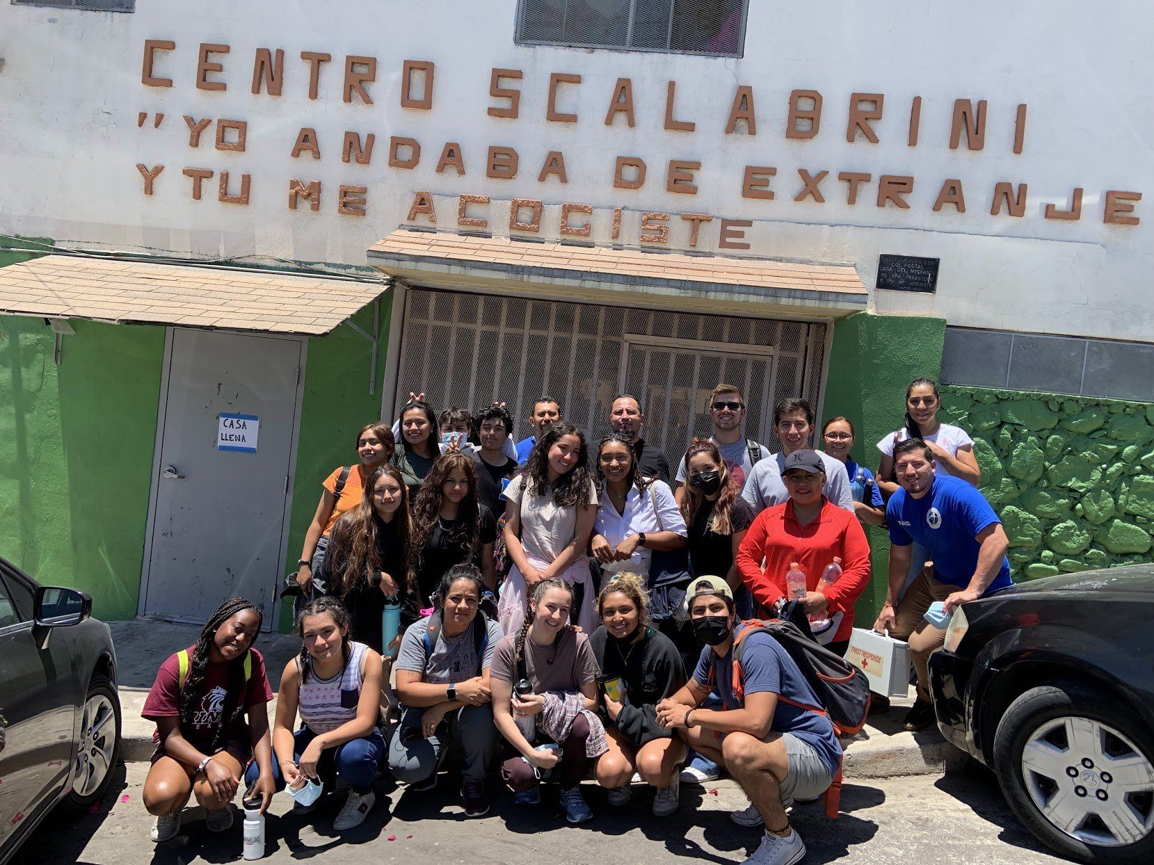 Group of people posing outside Centro Scalabrini, with a green wall and parked cars nearby.