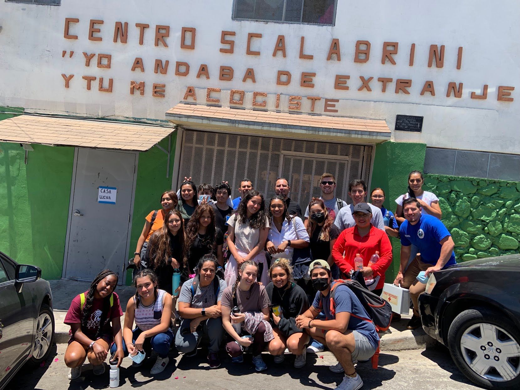 Group posing outside Centro Scalabrini entrance, with a green wall, parked cars, and a metal gate in front.