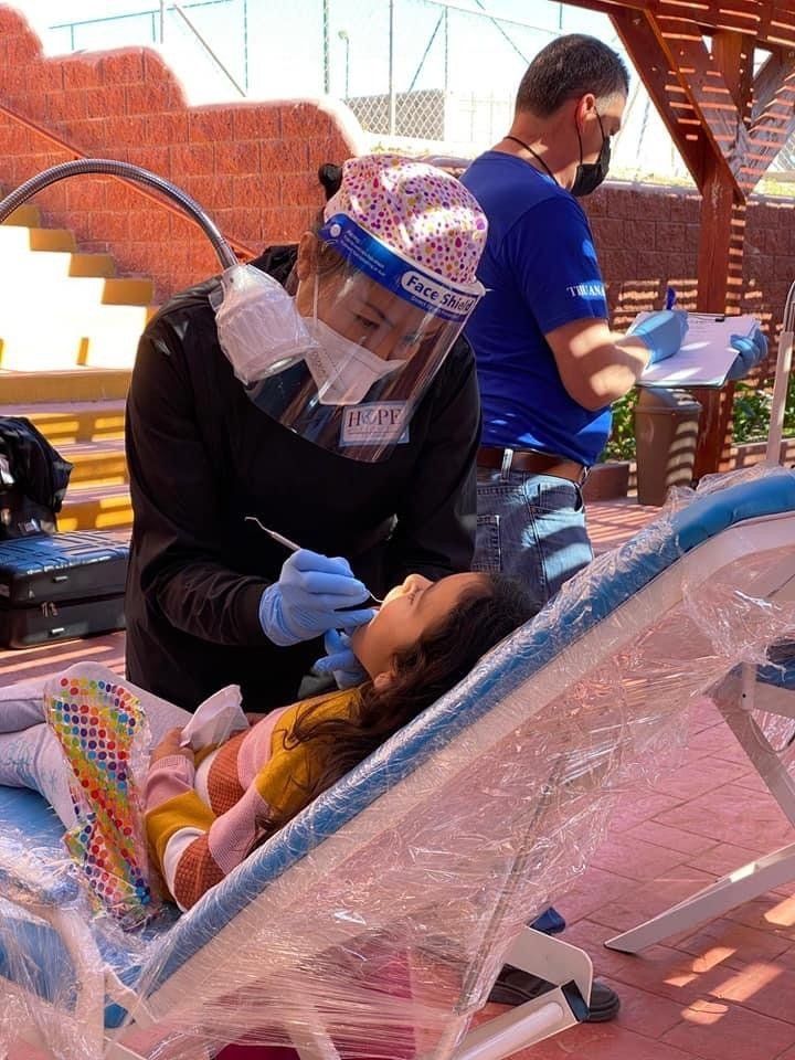 Healthcare worker in face shield and gloves examining a seated child outdoors behind clear plastic barrier