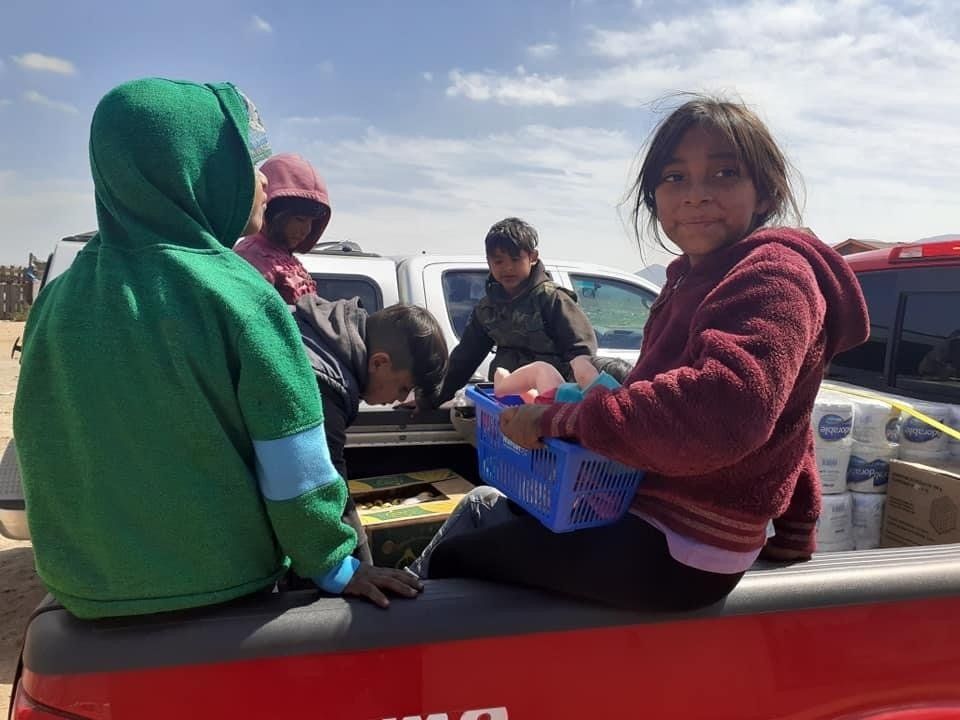 People sitting in the back of a pickup truck, with parked vehicles and a bright sky behind them.