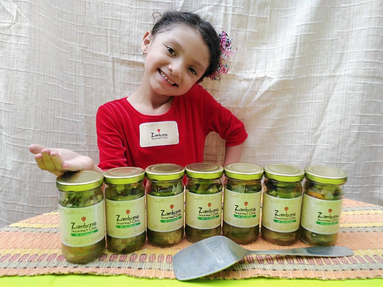 Child smiling behind a row of green-labeled jars on a table, holding one jar in each hand