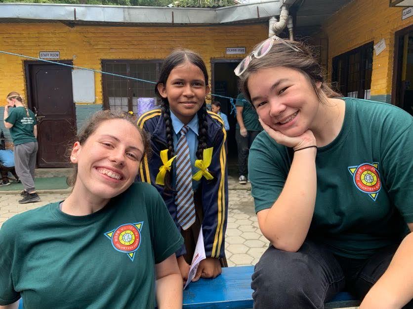 Three smiling students kneel in a school courtyard, wearing green shirts and a striped uniform.
