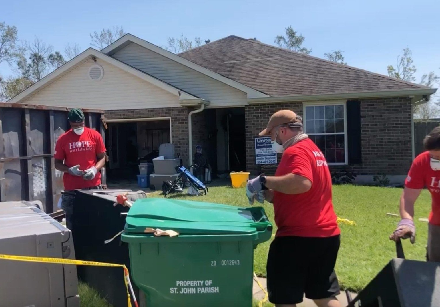 People in red shirts collect bins and items in a suburban driveway outside a house