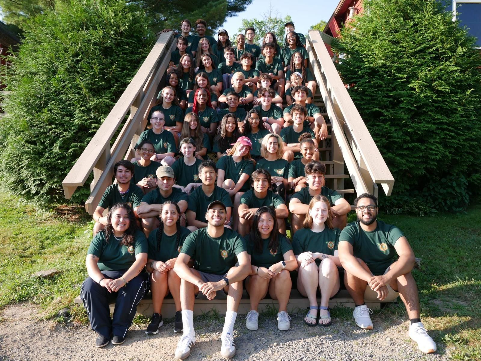 Large group posed on outdoor steps, wearing dark green shirts, in front of dense green trees.