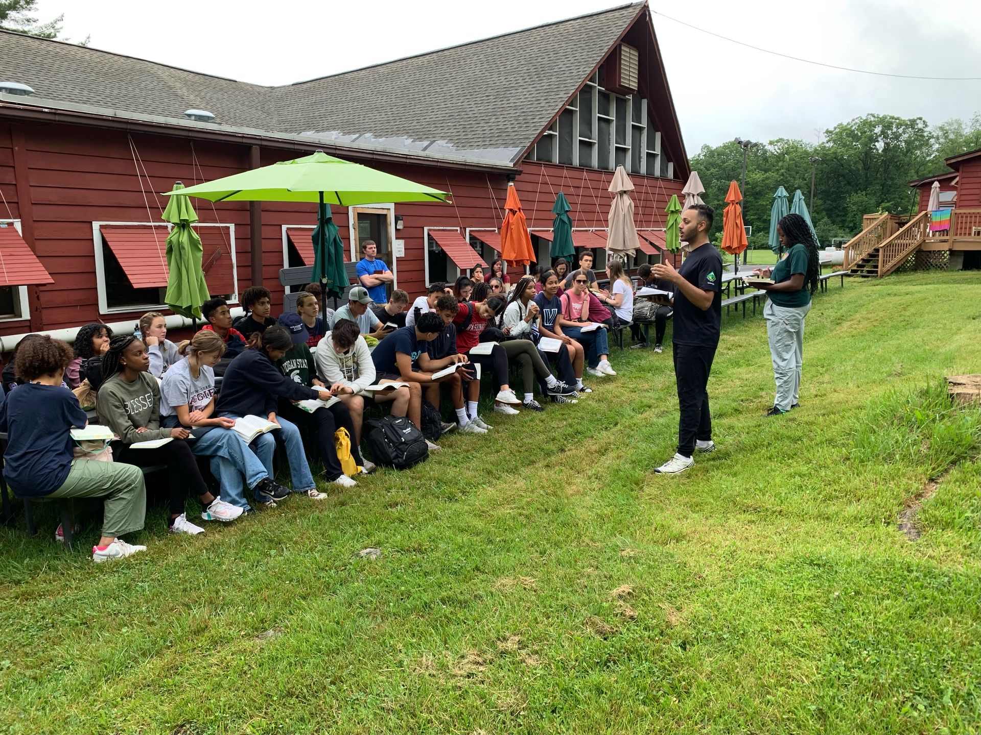 Outdoor group gathering by a red cabin, with people seated on benches and a speaker standing on the grass.