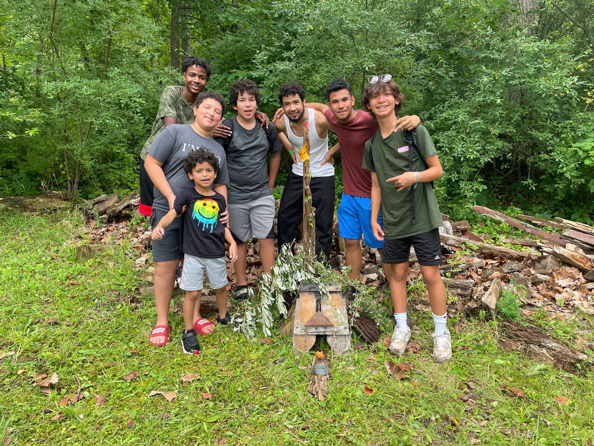Group of six people smiling outdoors in a lush forest, standing around a small grave marker.