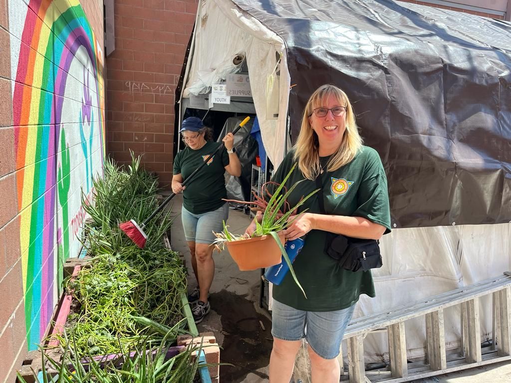 Two people carrying plants through a tented walkway beside a rainbow mural
