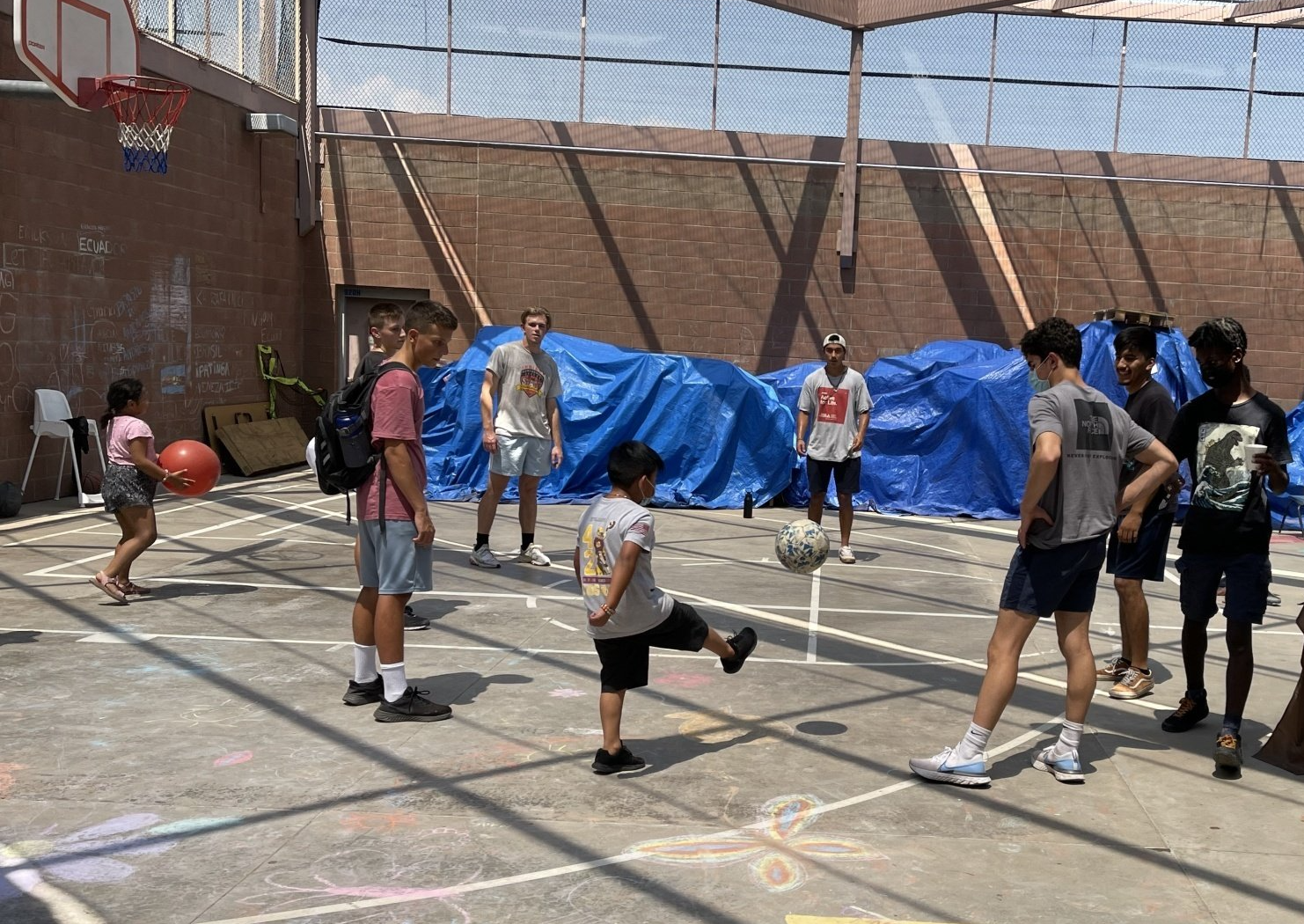 People playing basketball on an outdoor court near a brick wall and blue tarp.