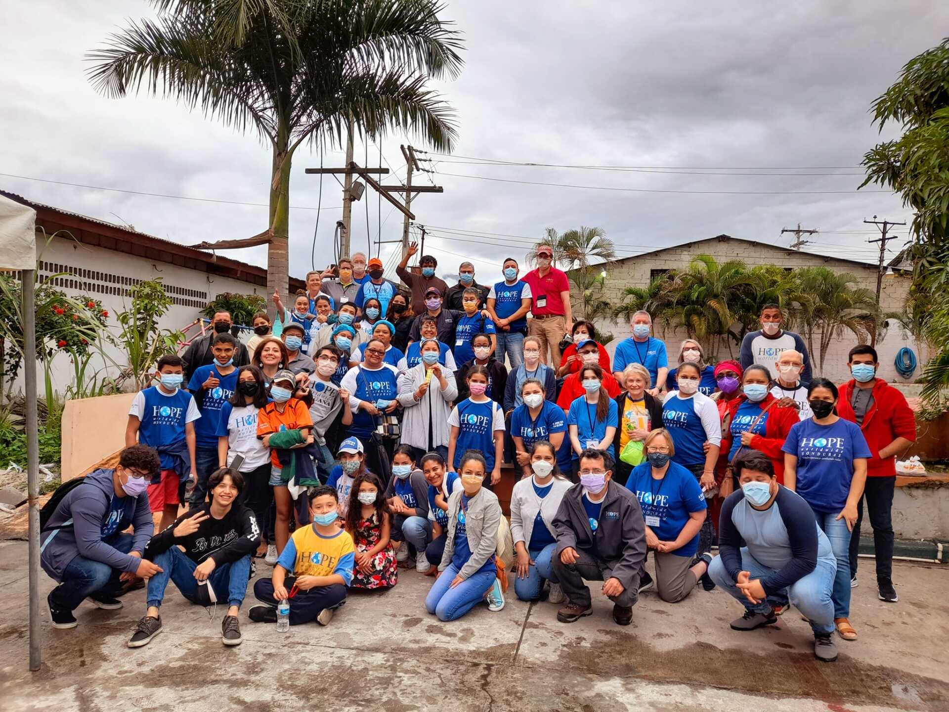 Large group posing outdoors in front of a house, many wearing blue shirts and face masks