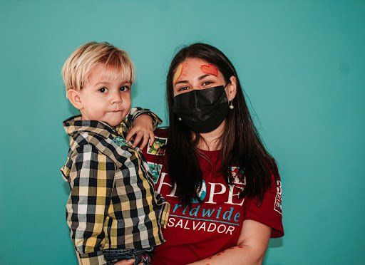 Woman in red shirt and black mask holding a child against a teal background