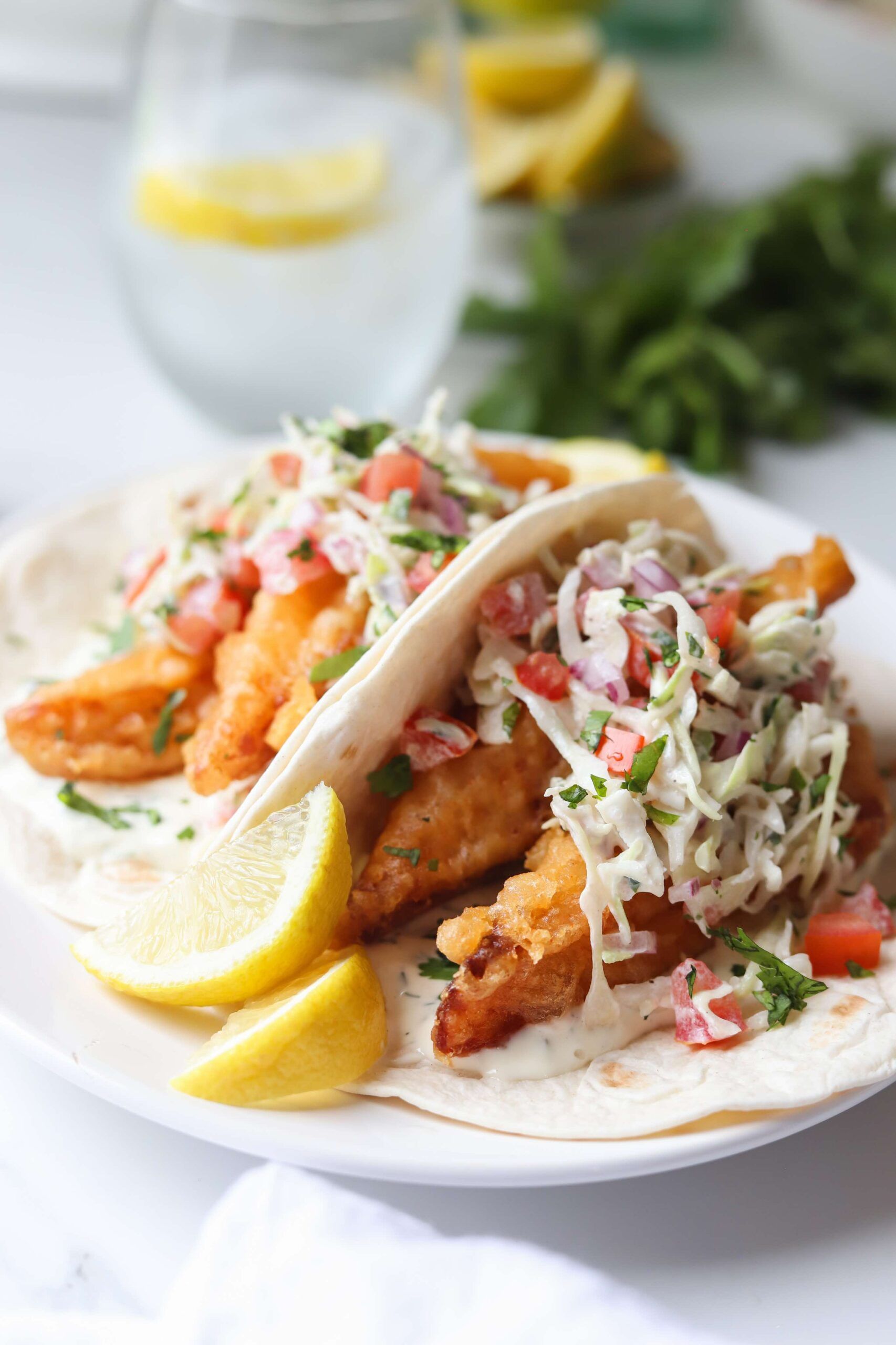 Two fish tacos on a white plate, topped with coleslaw and diced tomatoes, served with lemon wedges and a glass of water.