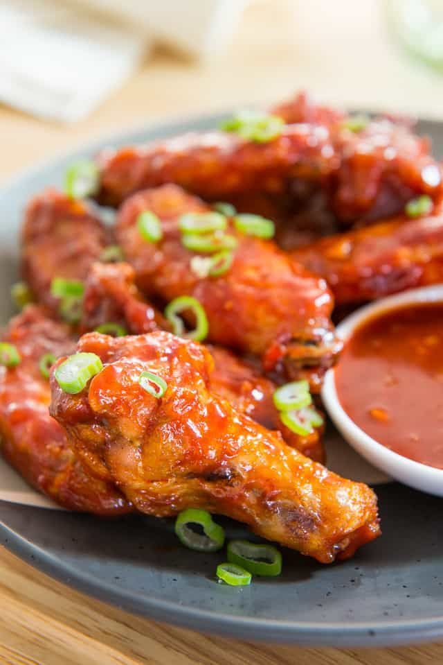 A plate of shiny, red-glazed chicken wings garnished with sliced green onions, served with a small bowl of dipping sauce.