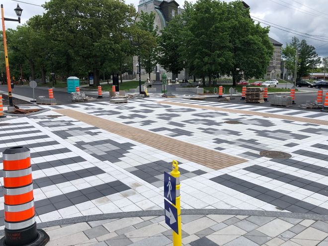 Passage piétonnier avec pavés à motifs et dalles podotactiles ; chantier de construction avec cônes orange et toilettes portables bleues.