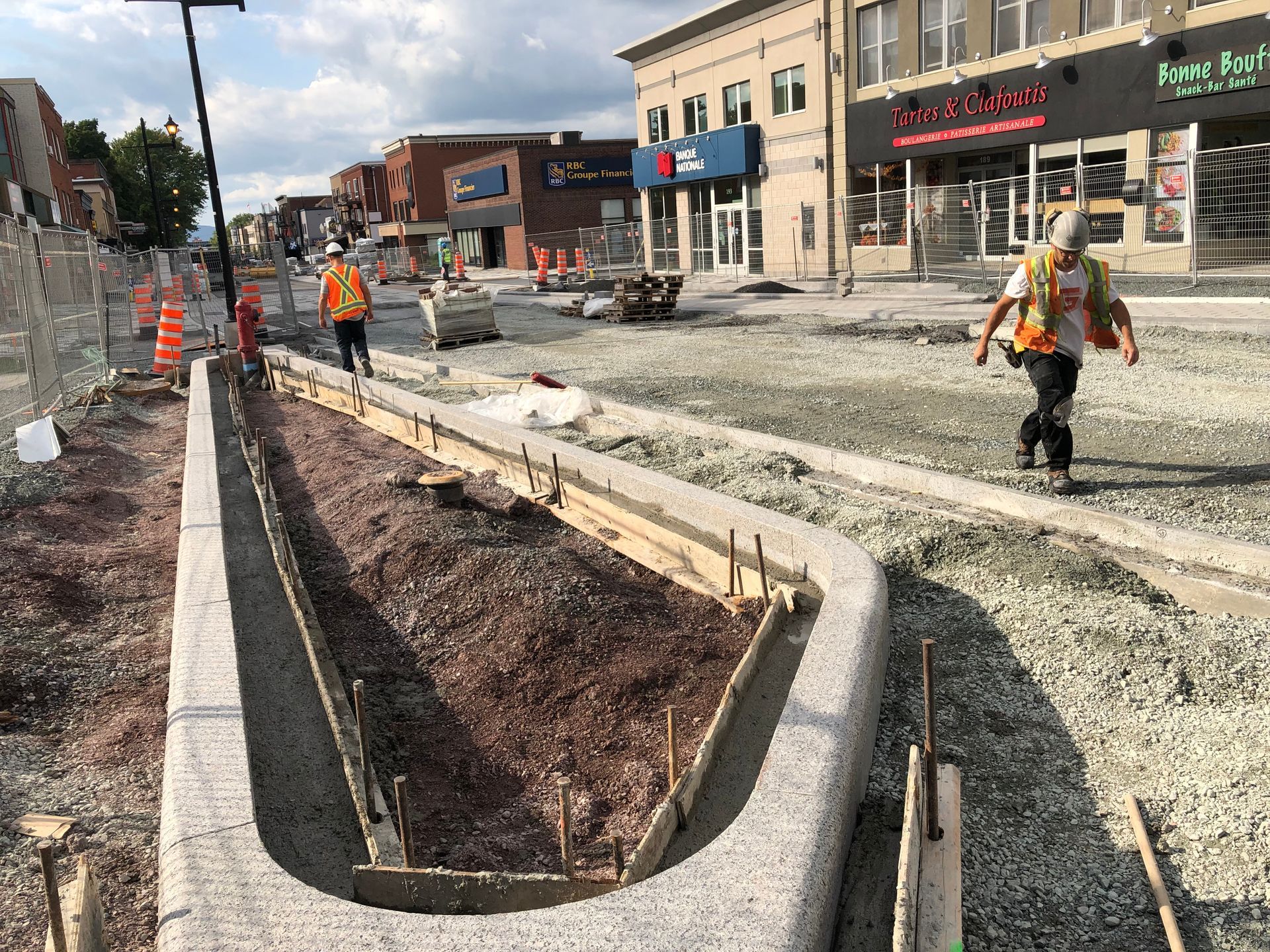 Travaux de voirie : Des ouvriers se trouvent près d’une jardinière en béton, avec du gravier, une route et des bâtiments en arrière-plan.