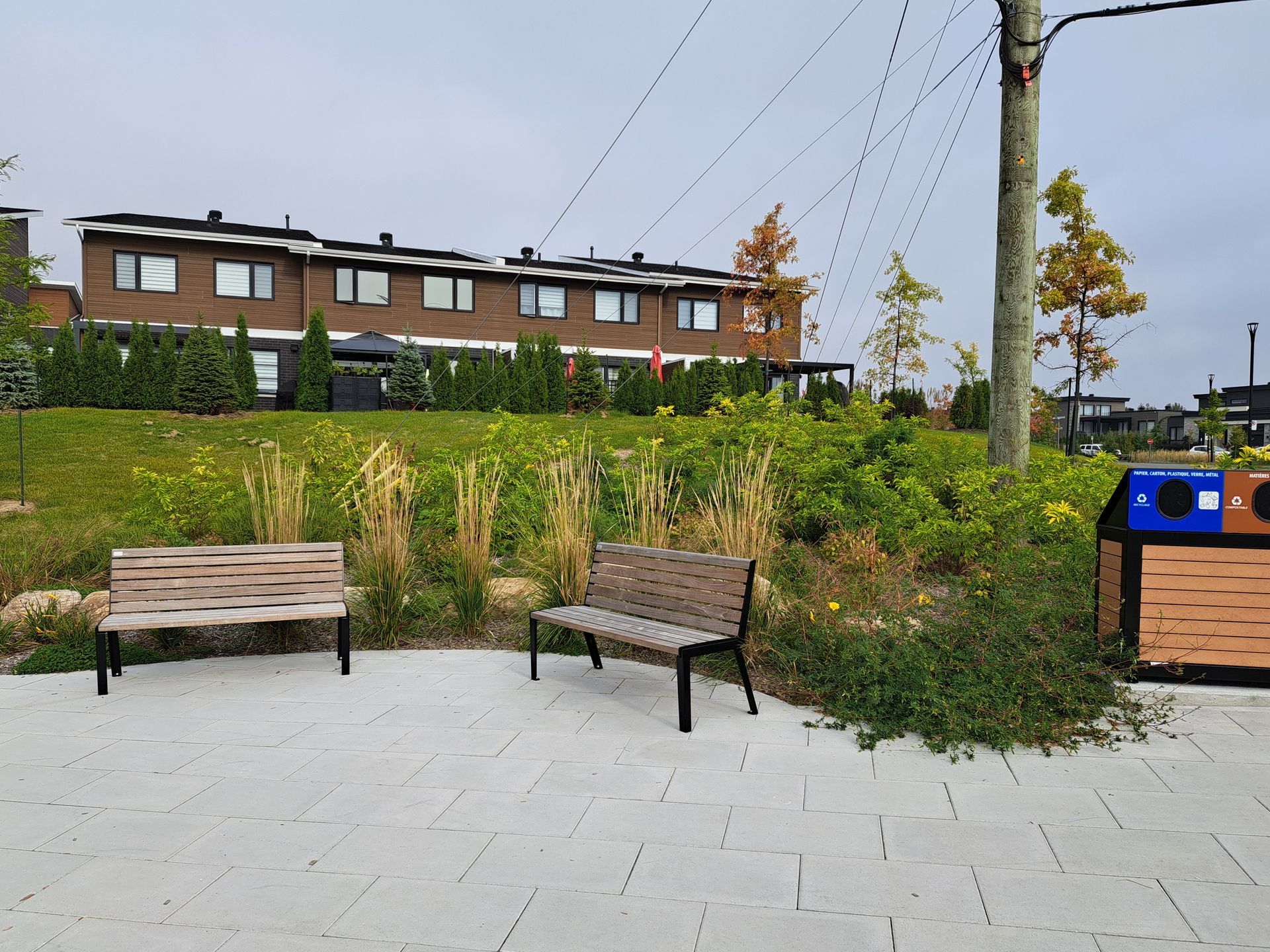 Deux bancs sur une esplanade pavée envahie par la végétation, face à des immeubles en briques de plusieurs étages.