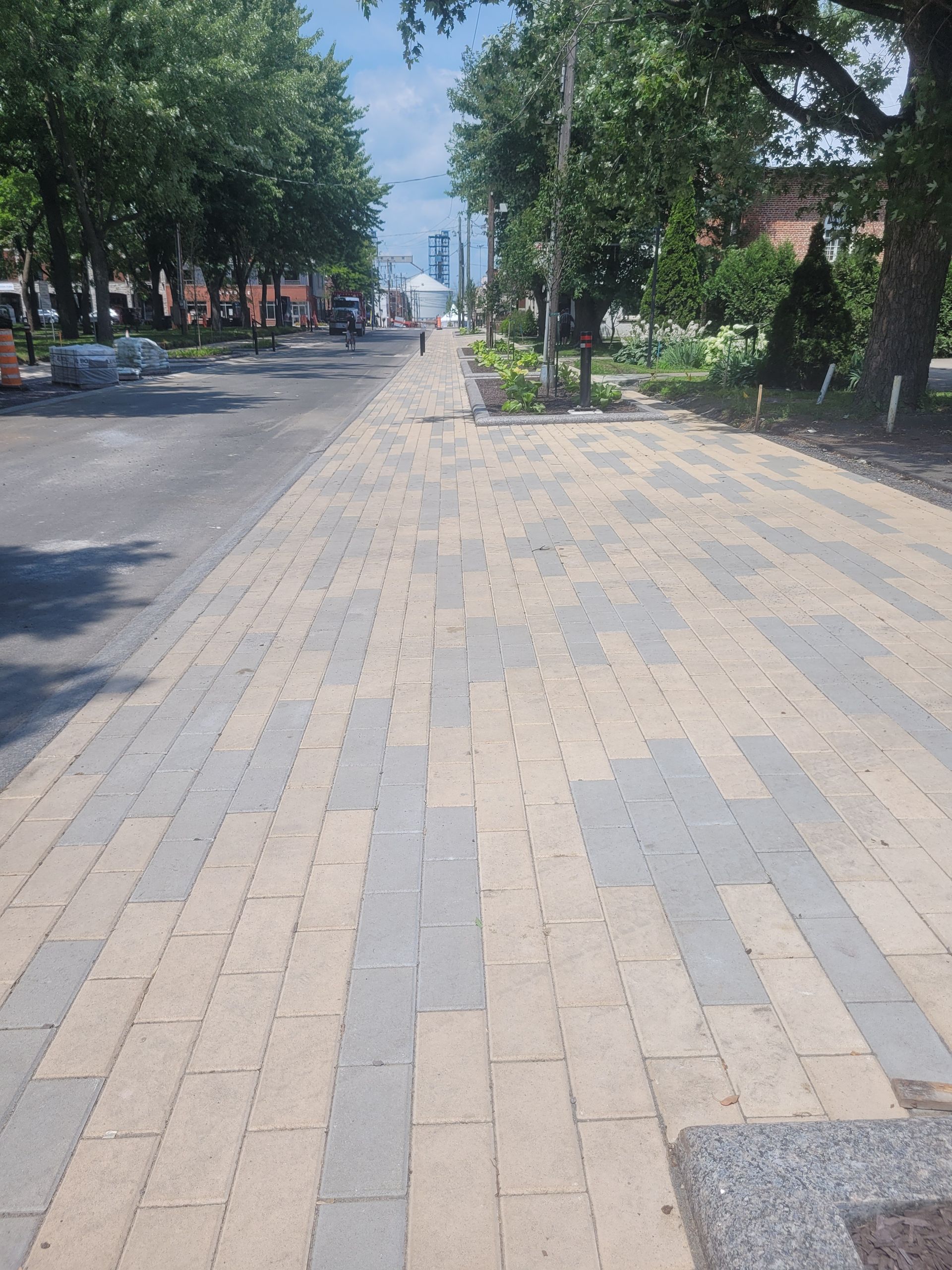 Un trottoir pavé de briques claires et foncées alternées mène à un espace arboré et verdoyant, semblable à un parc.