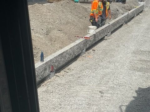 Des ouvriers installent une bordure de granit au bord d'un chemin de gravier. On aperçoit une personne en gilet orange et une autre, partiellement visible.