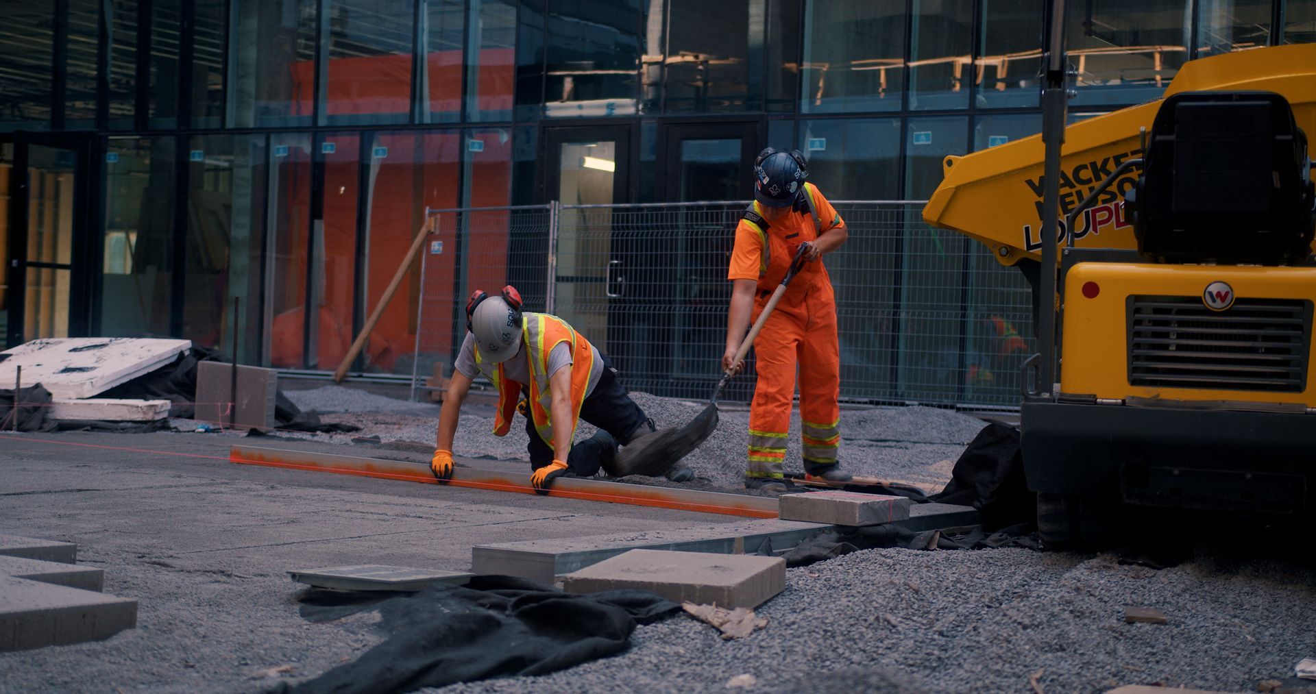 Deux ouvriers du bâtiment, vêtus de combinaisons et de gilets orange, travaillent sur un chantier, près d'un véhicule jaune.