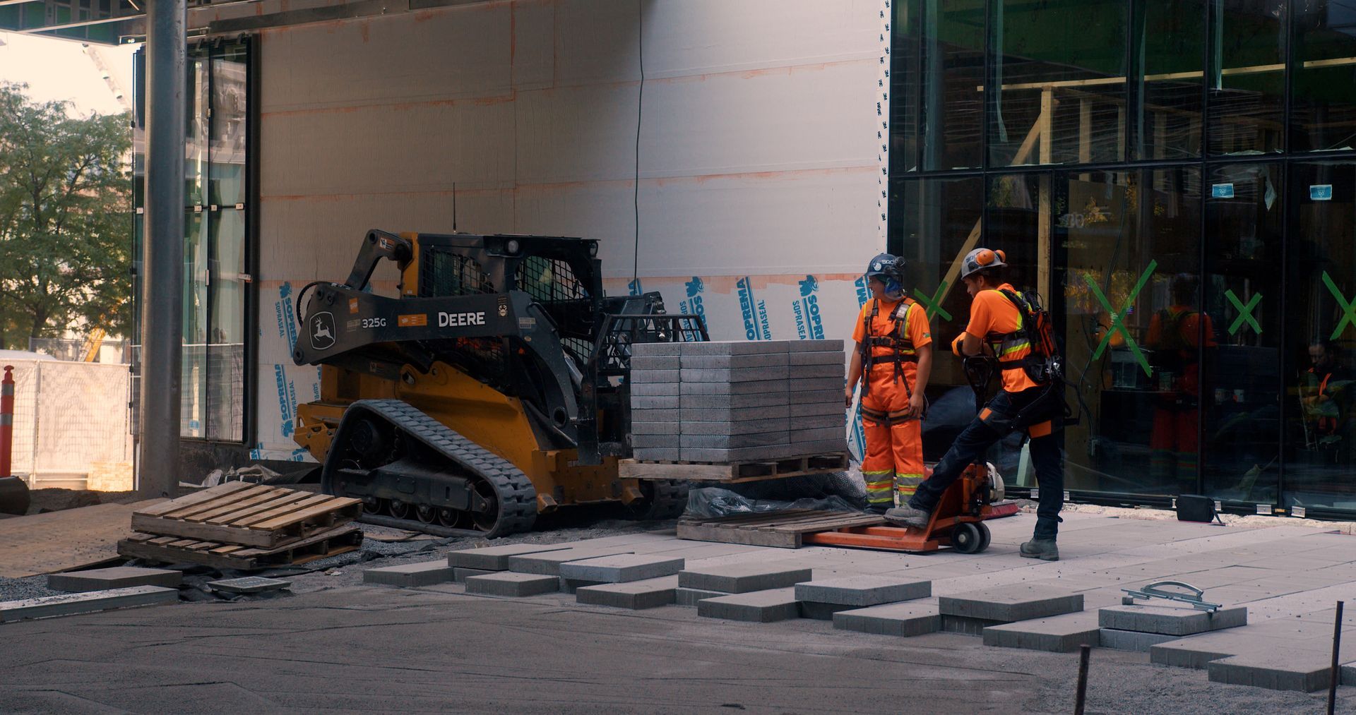 Chantier : Deux ouvriers en gilets orange près d'une chargeuse compacte et de paveurs.