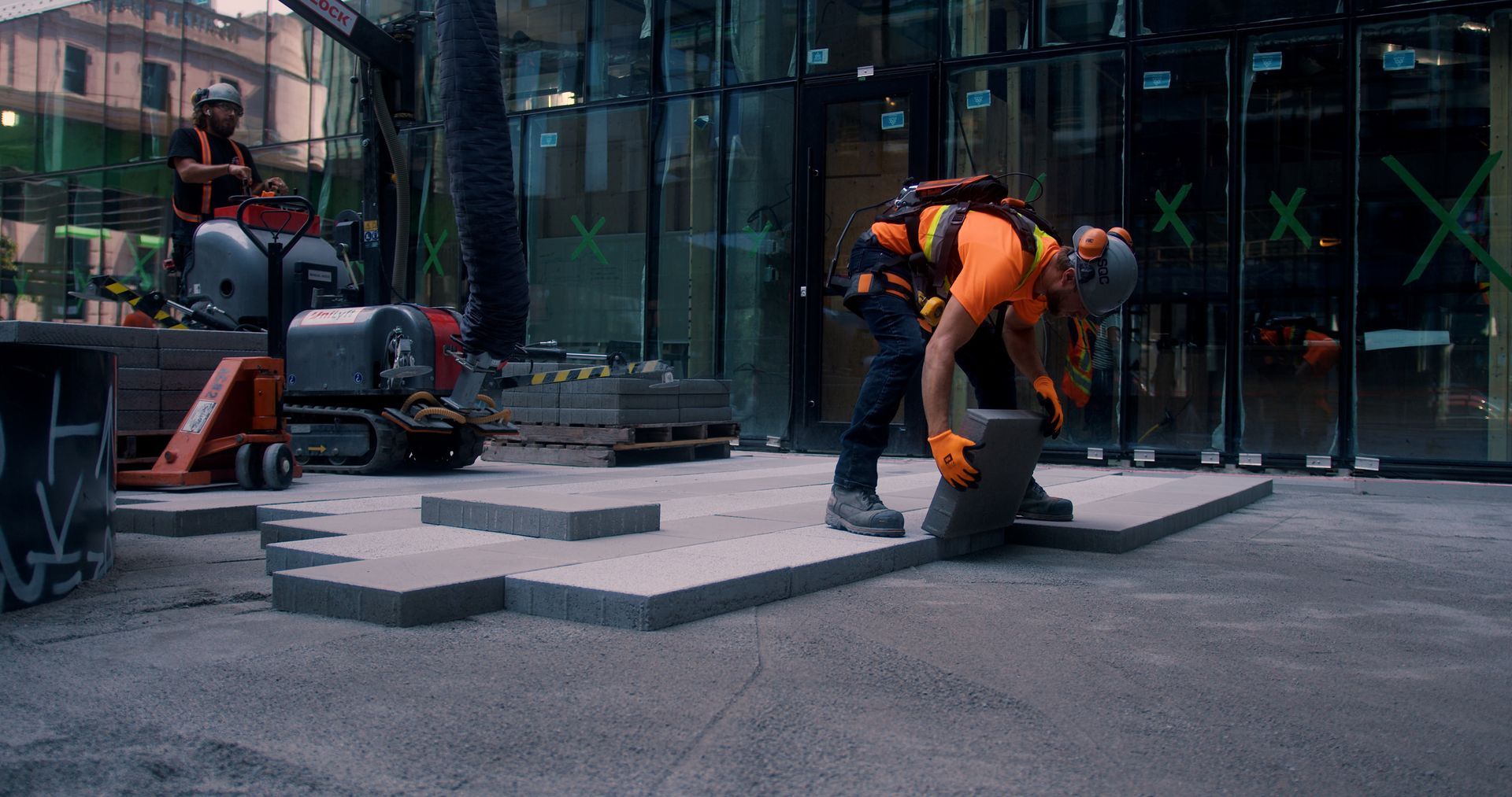 Ouvrier du bâtiment en gilet orange, posant une dalle grise. Un autre ouvrier est visible à l'arrière-plan. Chantier.