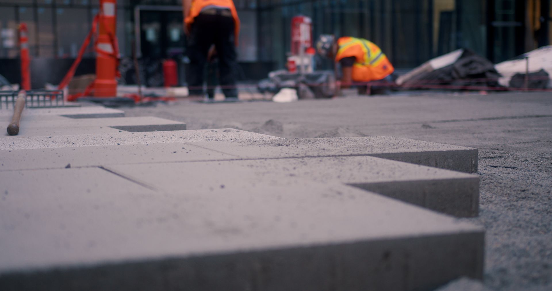 Des ouvriers du bâtiment vêtus de gilets orange travaillent sur une surface en béton à l'extérieur, l'un d'eux utilisant une scie.