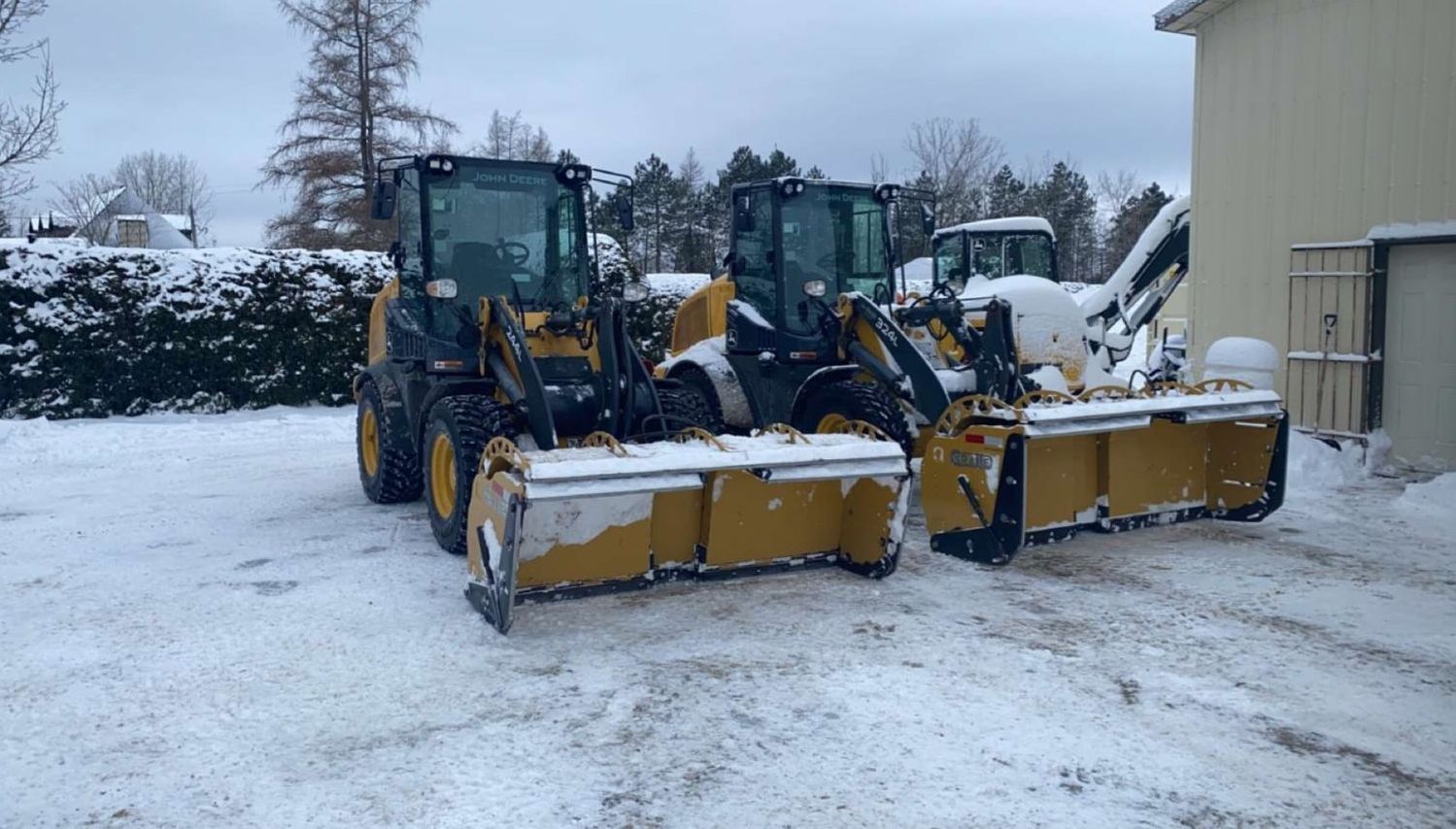 Deux chargeuses frontales jaunes équipées de souffleuses à neige sur une surface enneigée près d'un bâtiment.