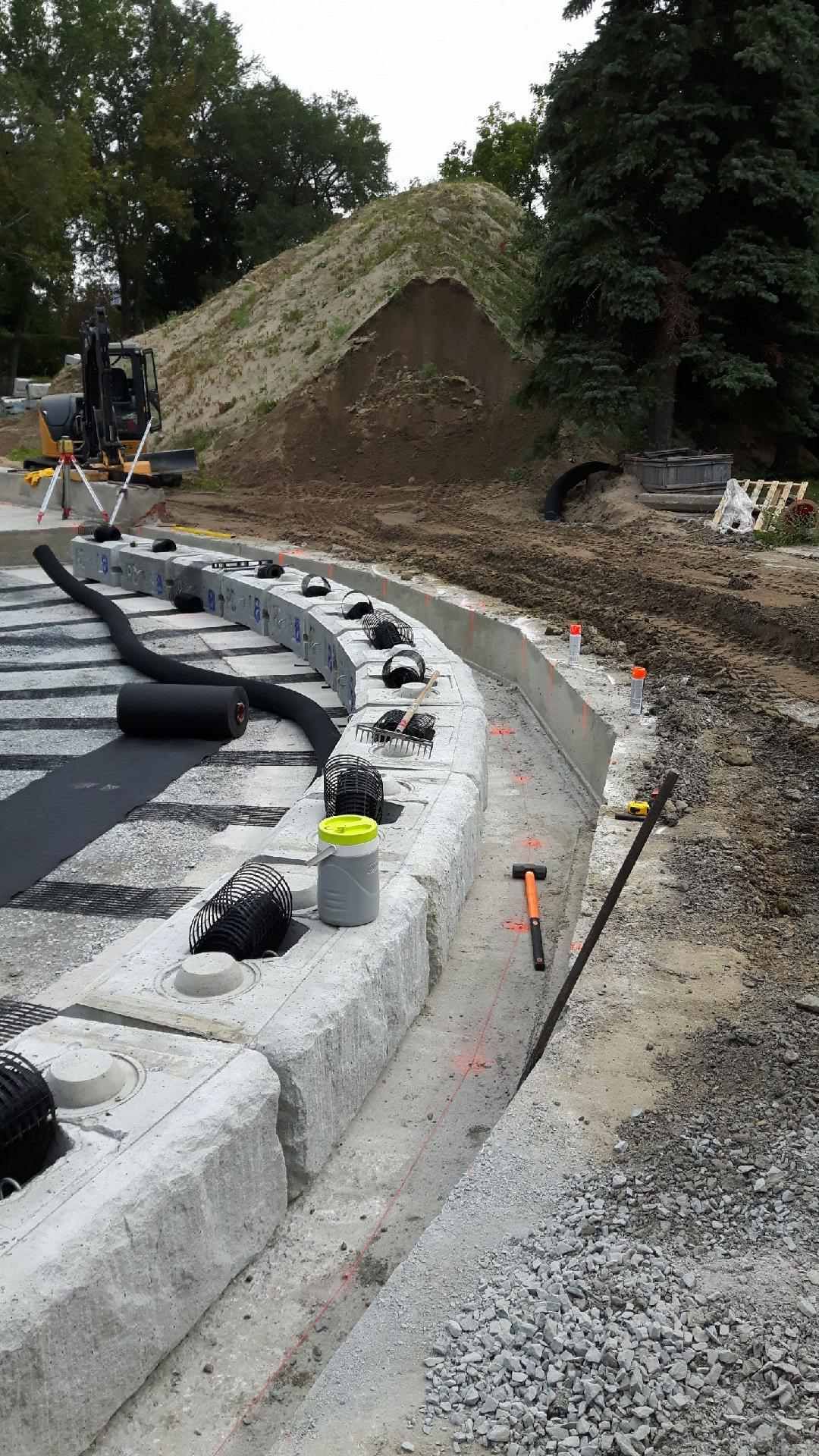 Chantier de construction avec mur de pierre incurvé en cours d'édification. Butte de terre, arbres en arrière-plan.