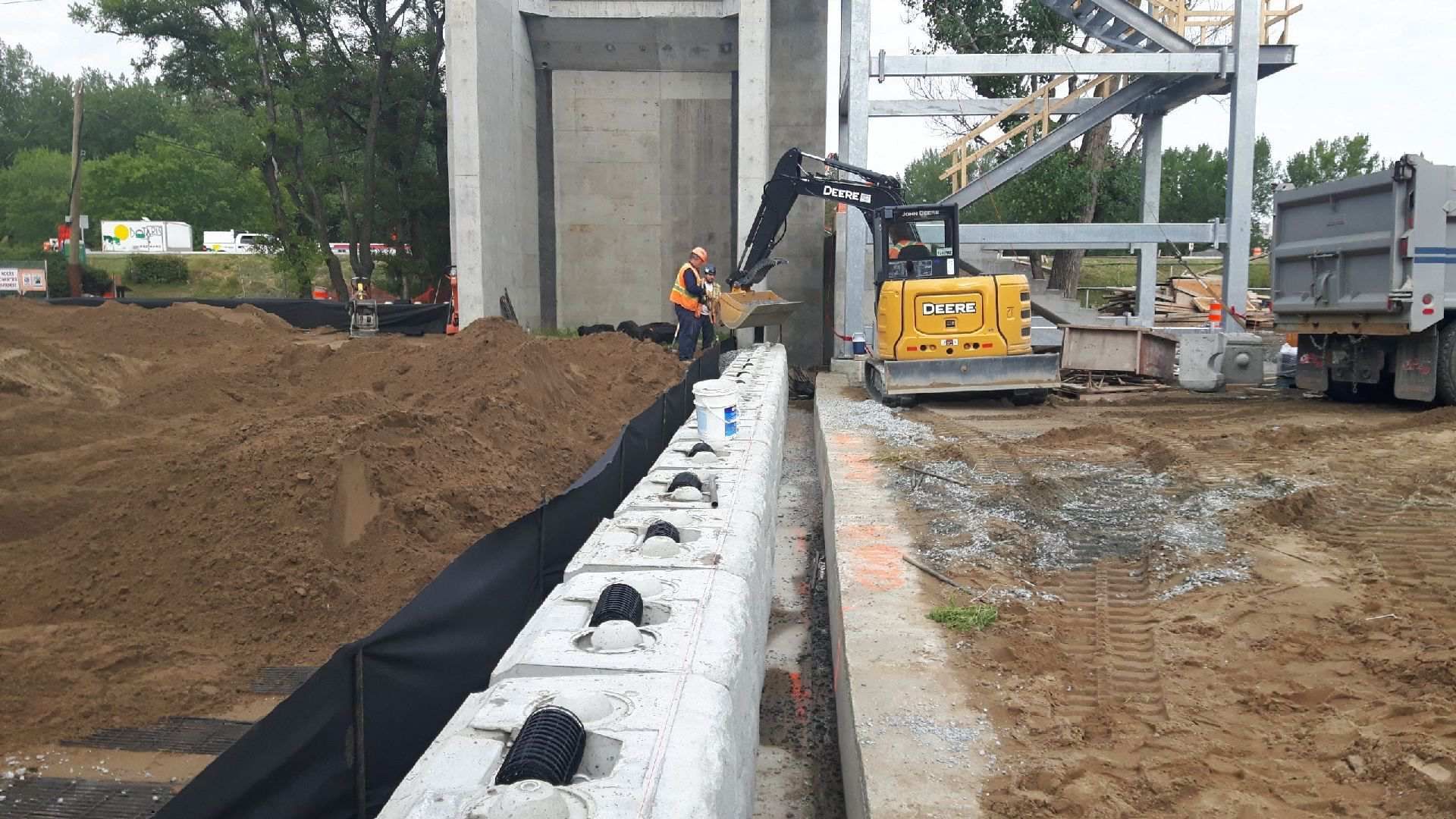 Chantier de construction : une excavatrice dépose de la terre contre un mur de soutènement ; un ouvrier est présent.