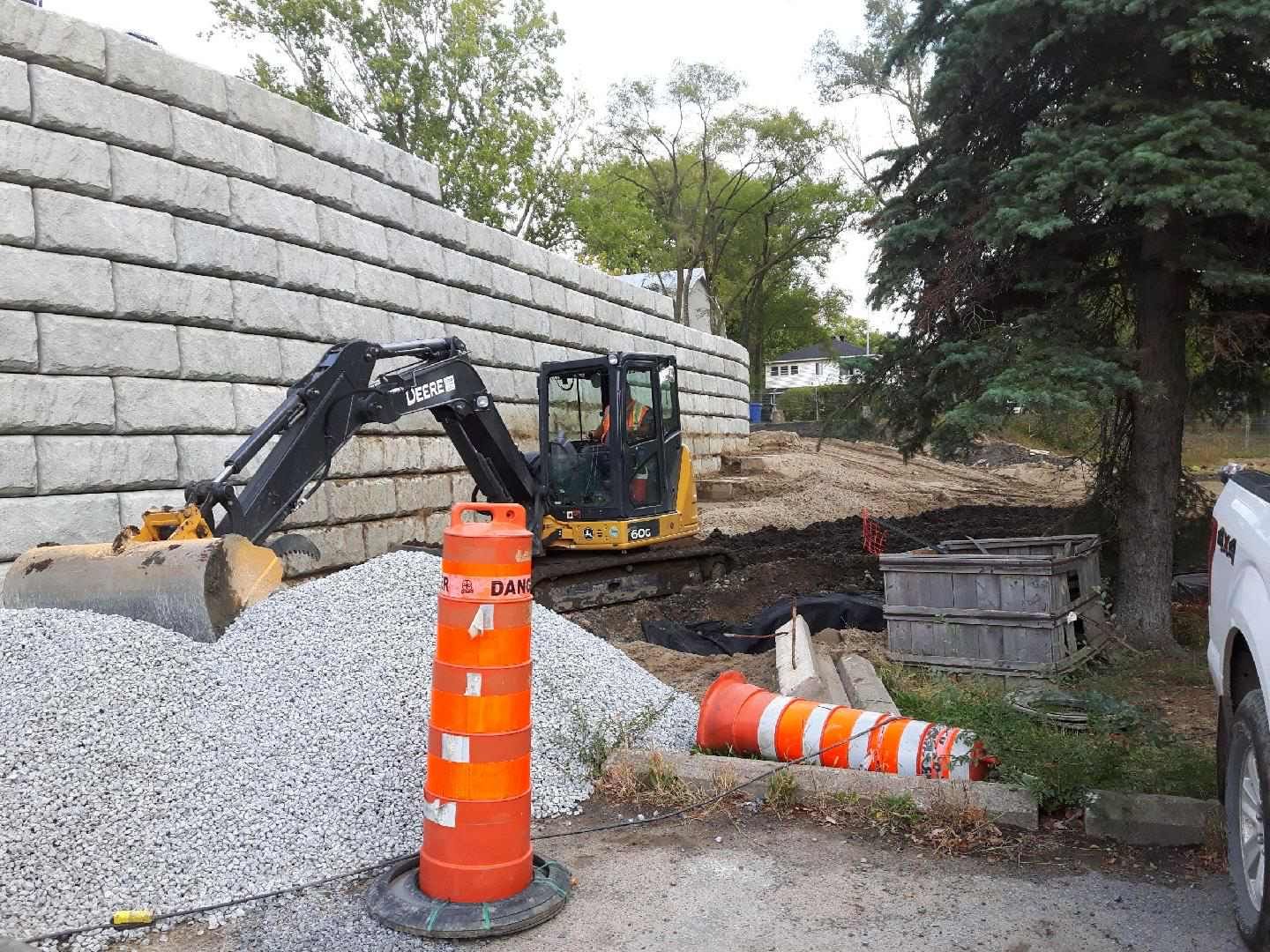 Une excavatrice travaille sur un chantier. Mur de soutènement gris, cônes de signalisation orange et tas de gravier.