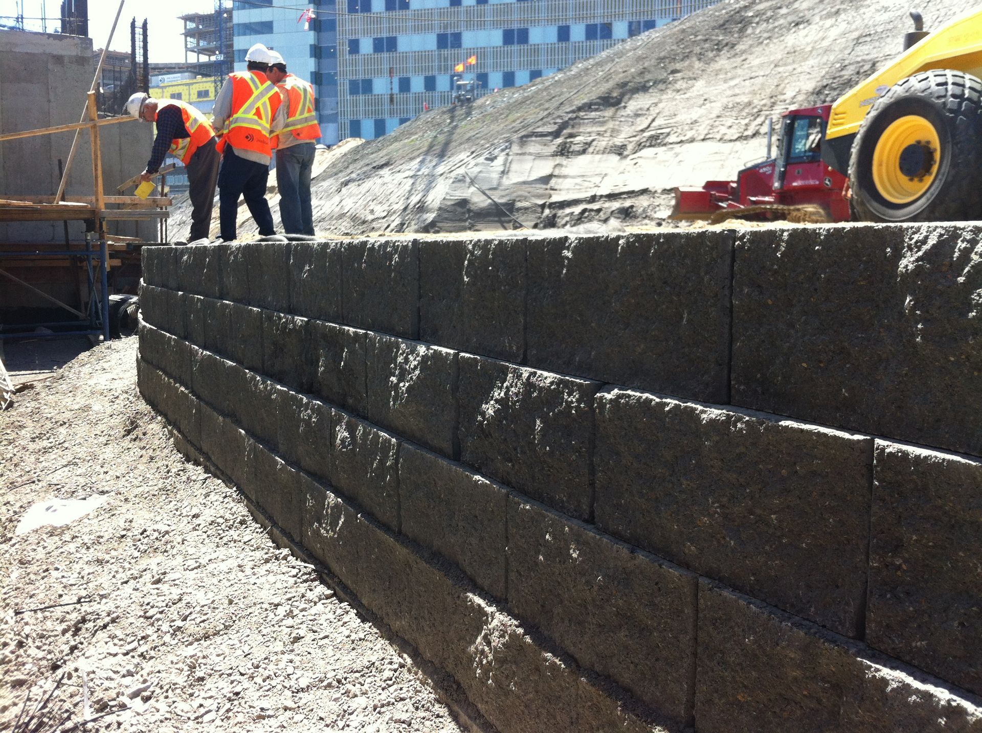 Des ouvriers inspectent un mur de soutènement en blocs gris foncé. Un bulldozer jaune se trouve à proximité.