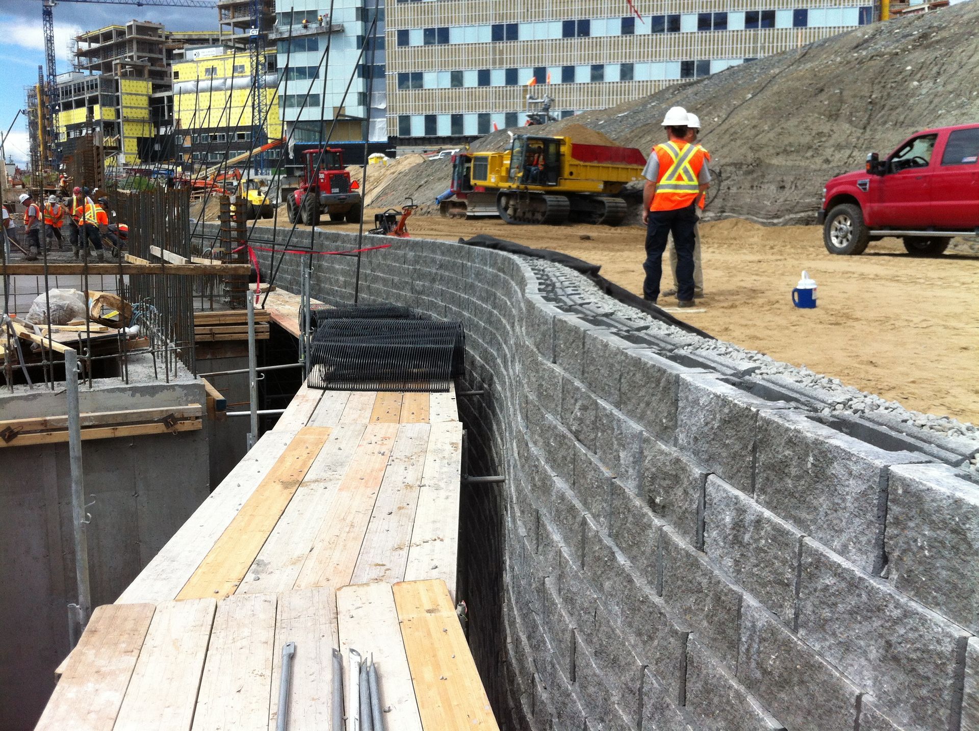 Chantier de construction avec ouvriers, mur de soutènement, engins de chantier et camion.