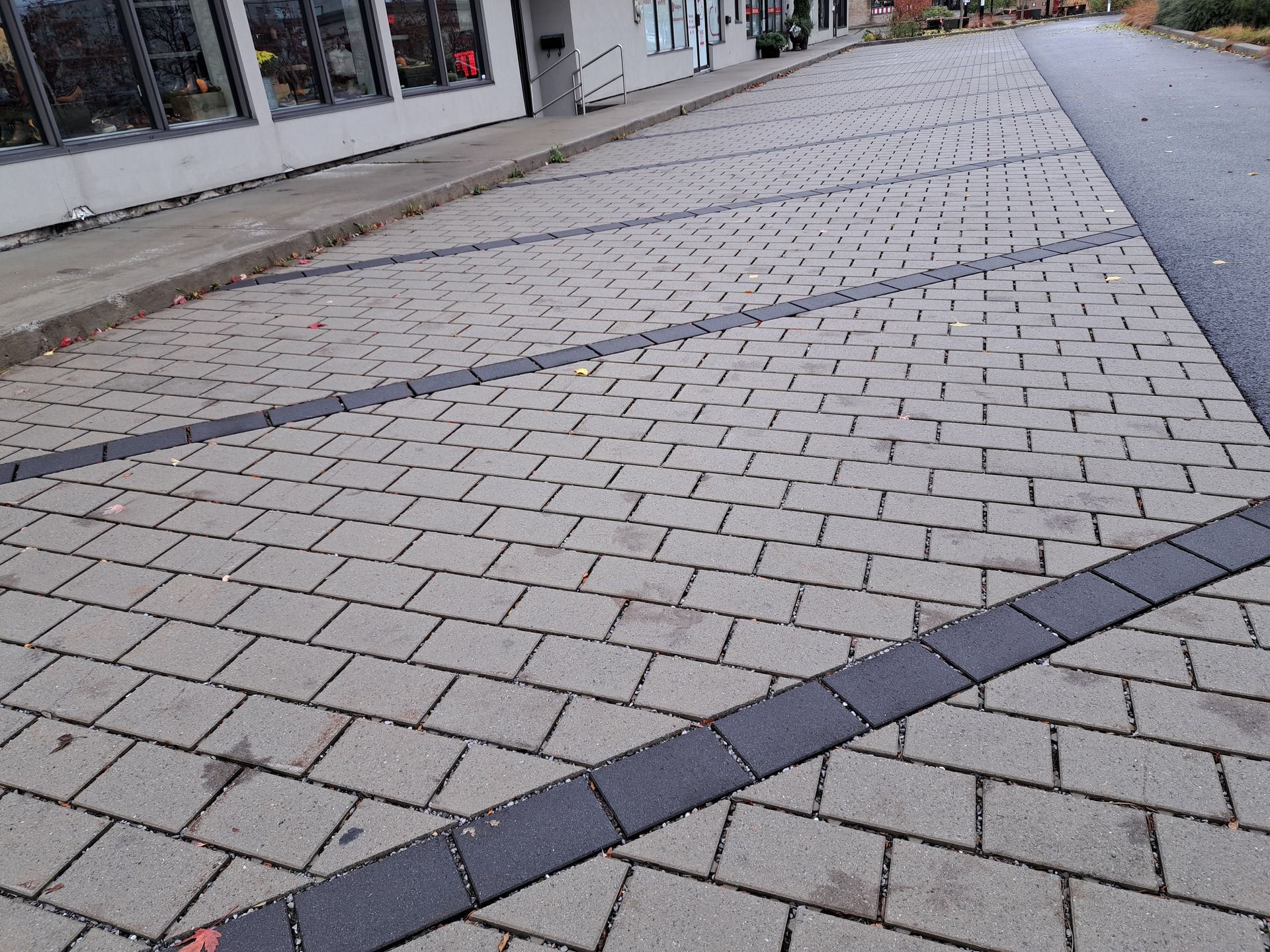 Zone pavée de briques aux bordures gris foncé, jouxtant une rue et un bâtiment.