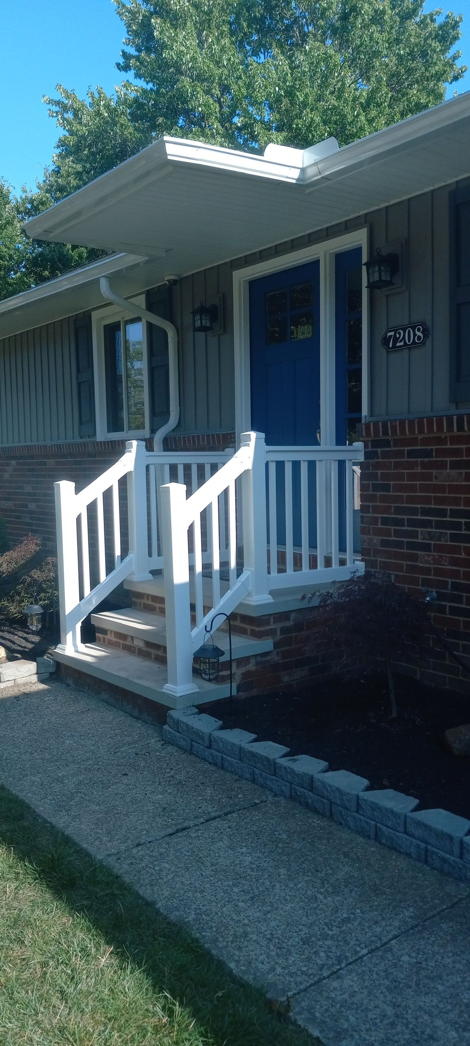 Front porch of a house with blue door, white railing, and brick steps in daylight