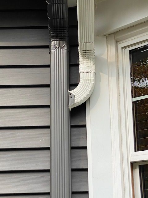 House siding with a gray downspout and white gutter beside a window