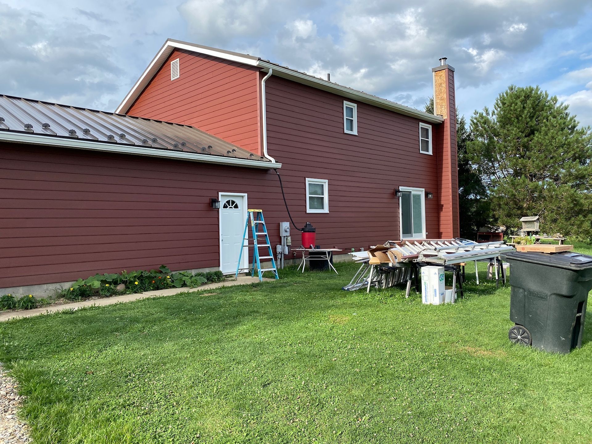 Red house with white trim, grassy yard, and patio furniture beside a utility pole under a cloudy sky