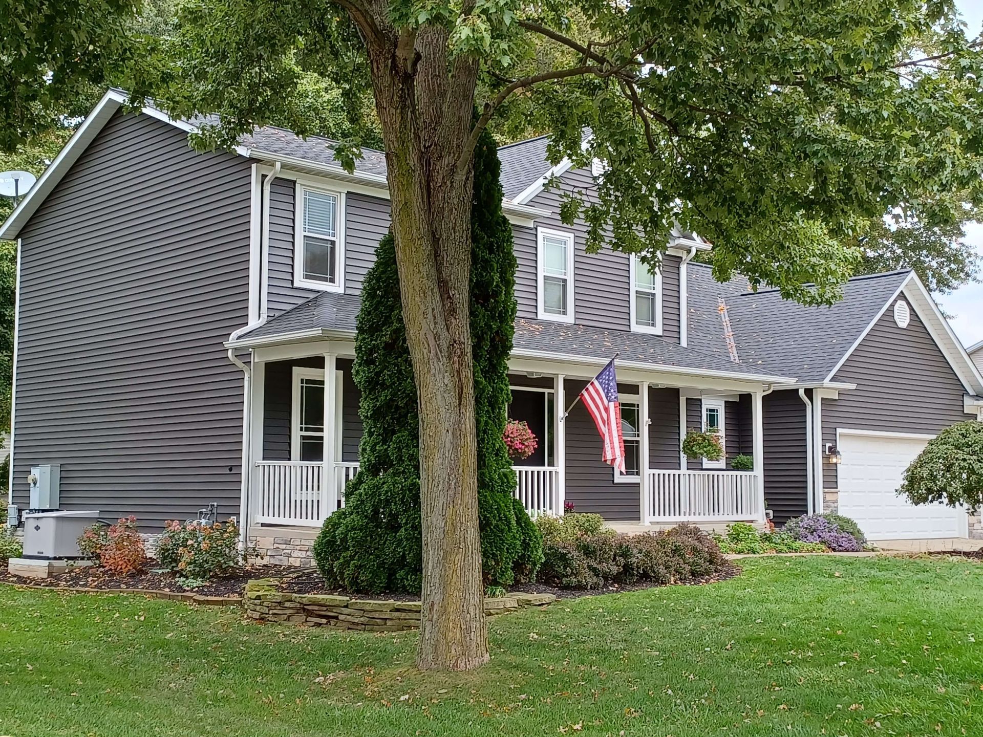 Gray suburban house with white trim, a front porch, American flag, and large trees in the yard.