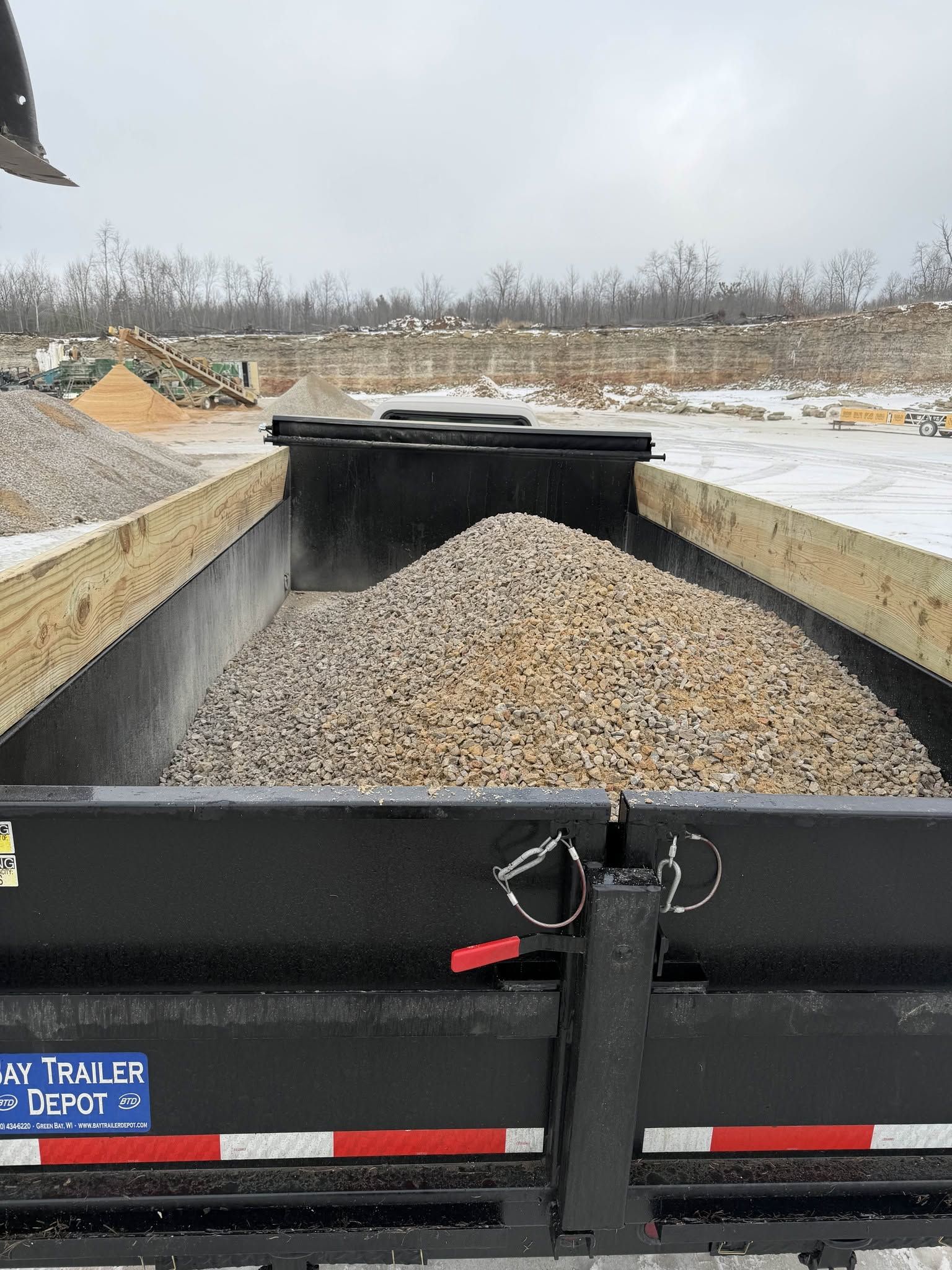 Black dump trailer filled with gravel at a construction site, viewed from the rear.