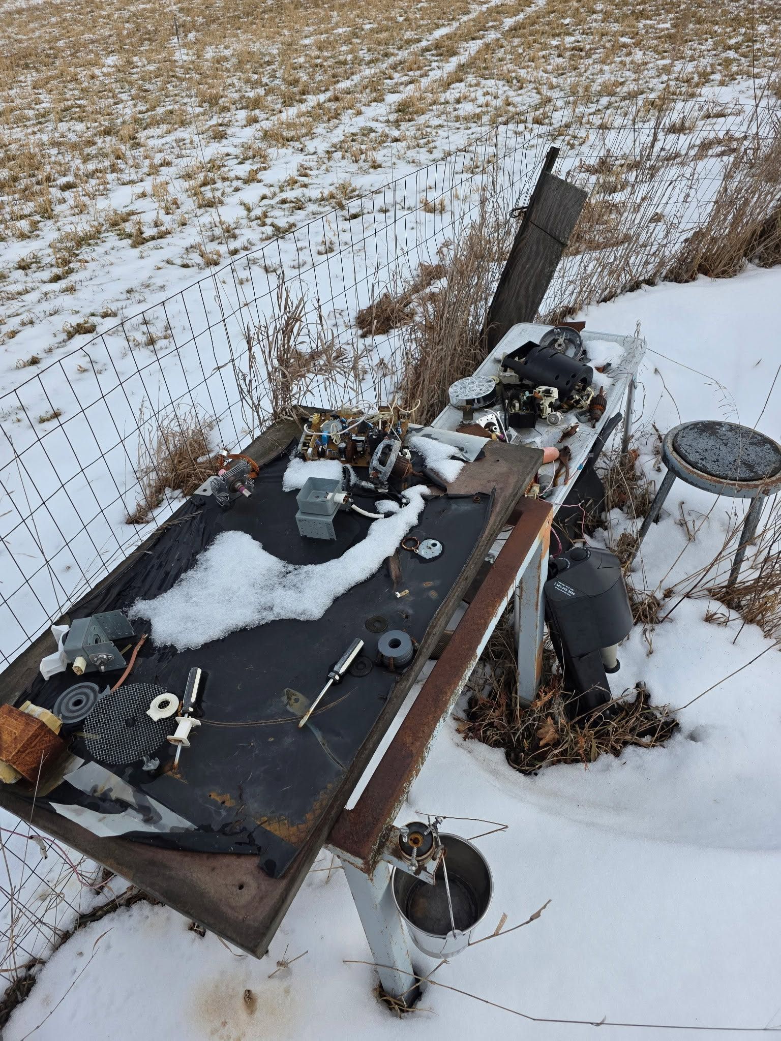 Overturned black trailer in snow beside a road, with scattered debris and a damaged round table.