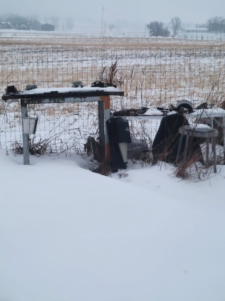 Snow-covered rural yard with a wooden table and scattered objects beside a fenced field in winter.