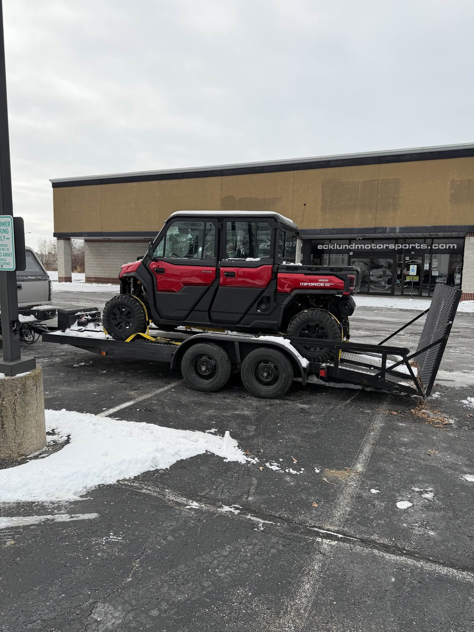 Red off-road side-by-side on a black utility trailer in a snowy parking lot.