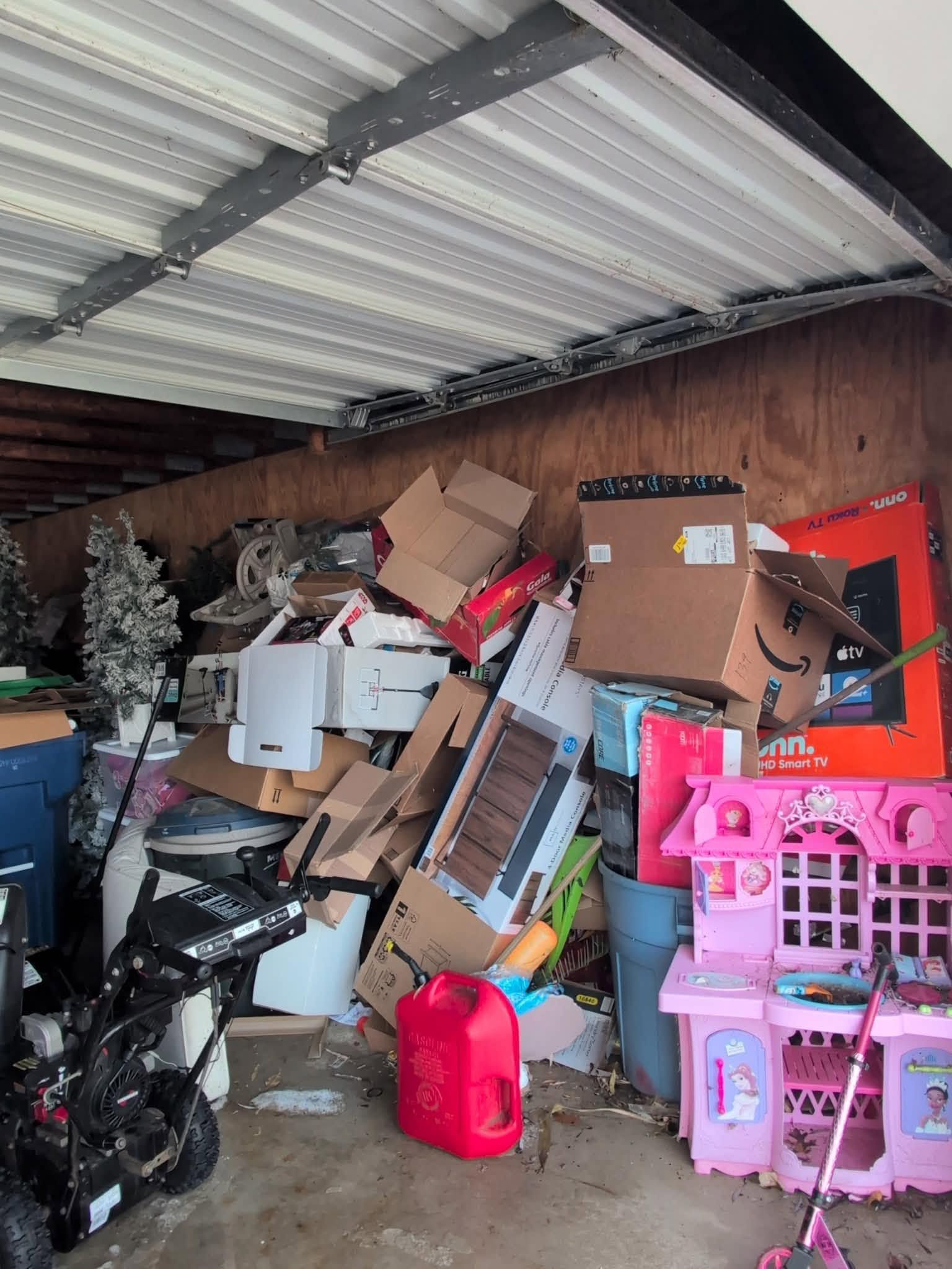 Cluttered garage with stacked boxes, furniture, appliances, and pink plastic storage bins under a corrugated roof