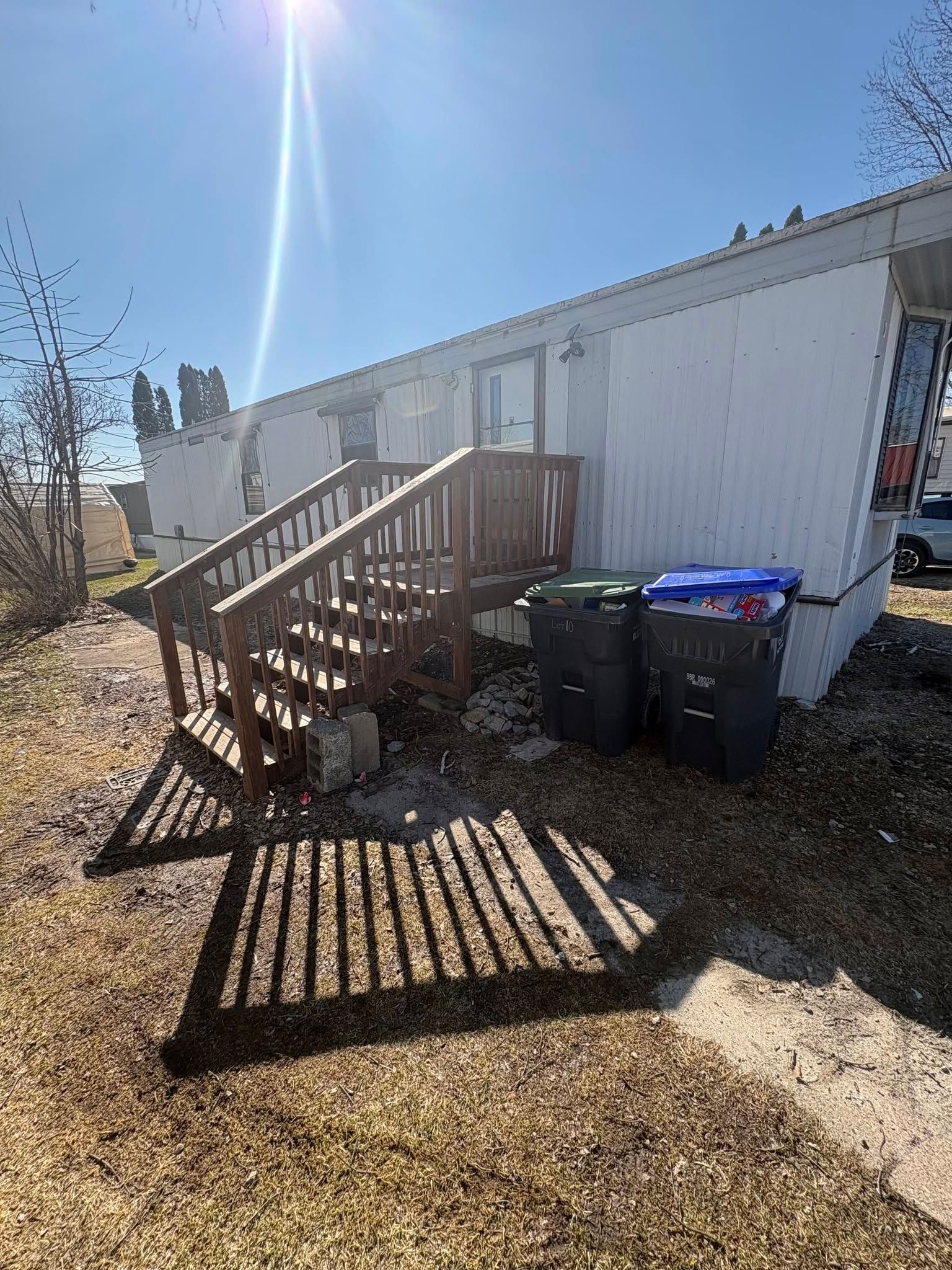 Mobile home exterior with wooden steps, trash bins, and long shadows in bright sunlight