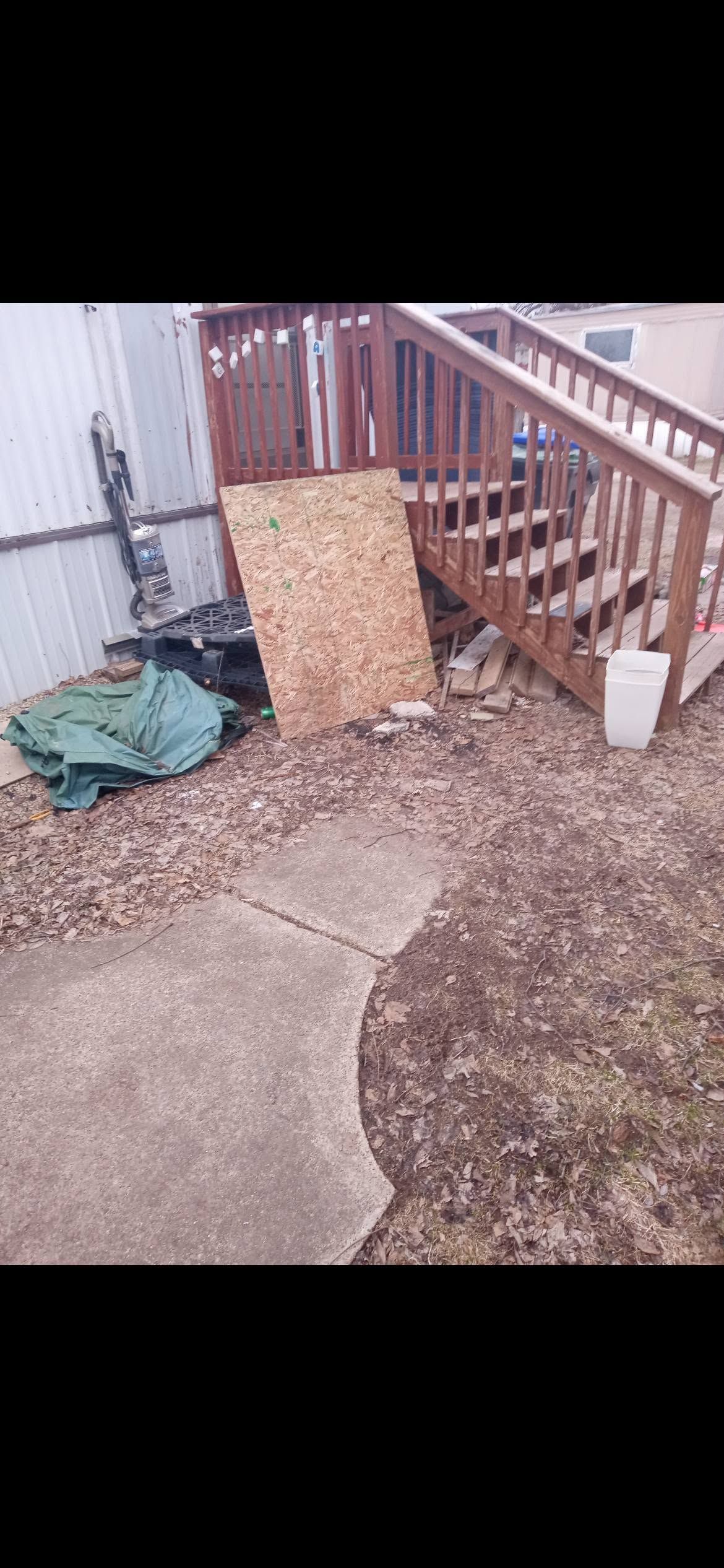 A small backyard patio with a wooden fence, leaning board, and a green tarp on the ground.