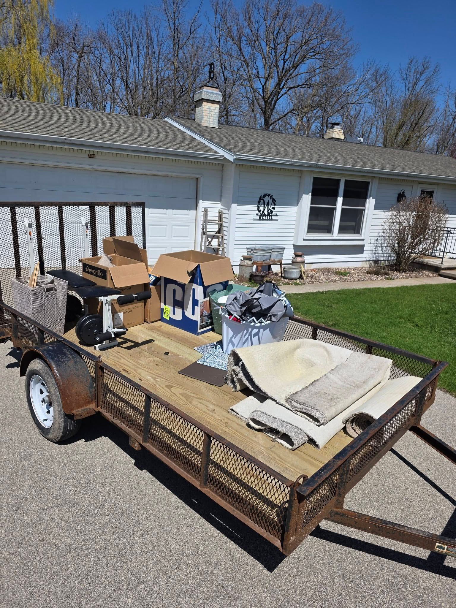 Trailer loaded with boxes, tools, and materials parked outside a white house on a sunny day