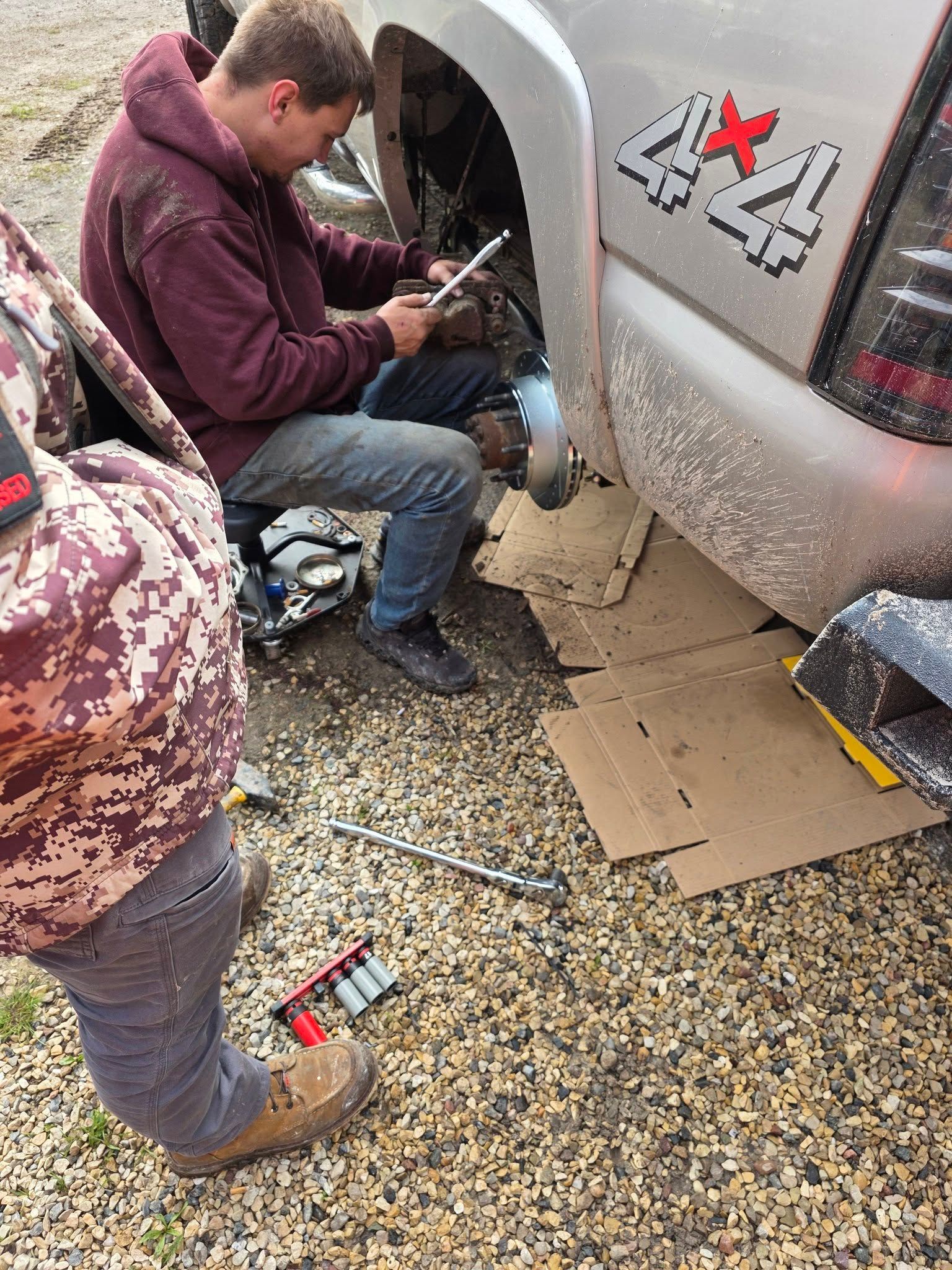 Two people work on a damaged white car’s wheel area on gravel, with tools and parts on the ground.